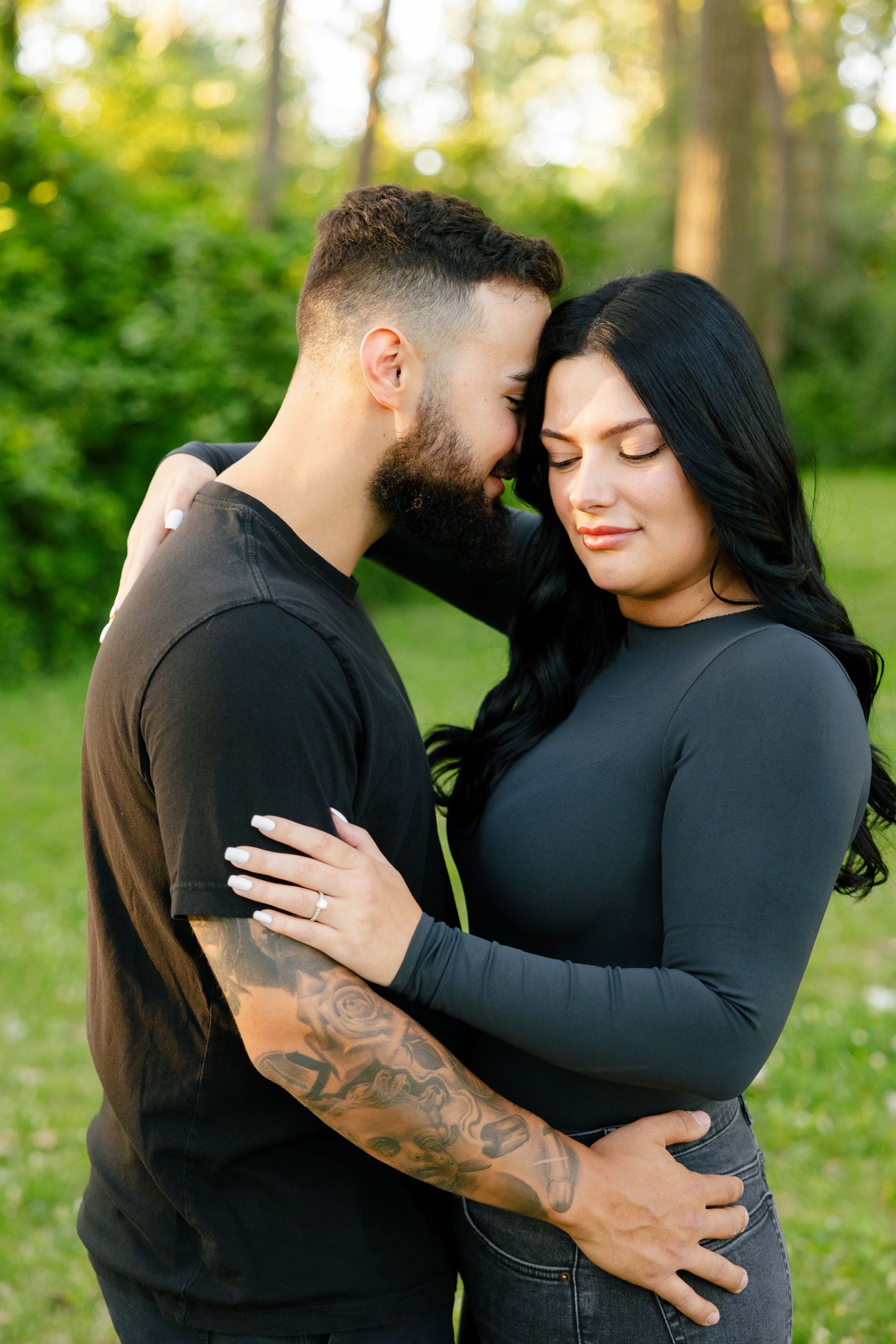 A couple in matching dark outfits embraces in a serene forest setting with soft lighting.