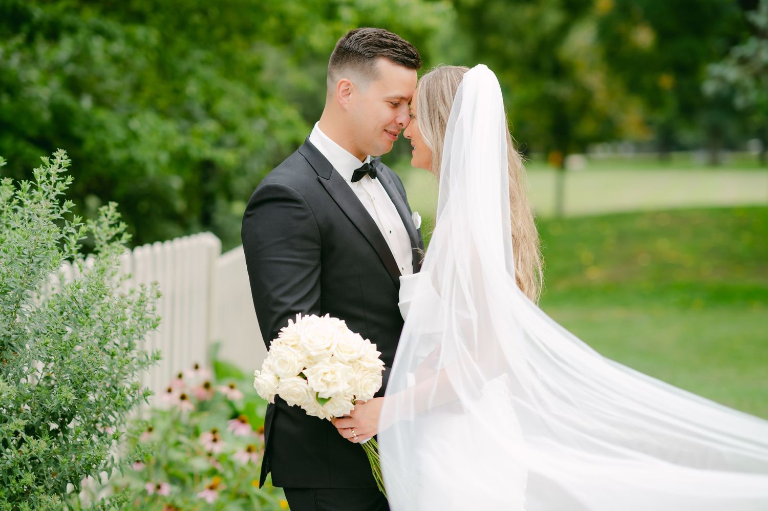 A couple poses in a garden setting with white roses and a flowing veil.