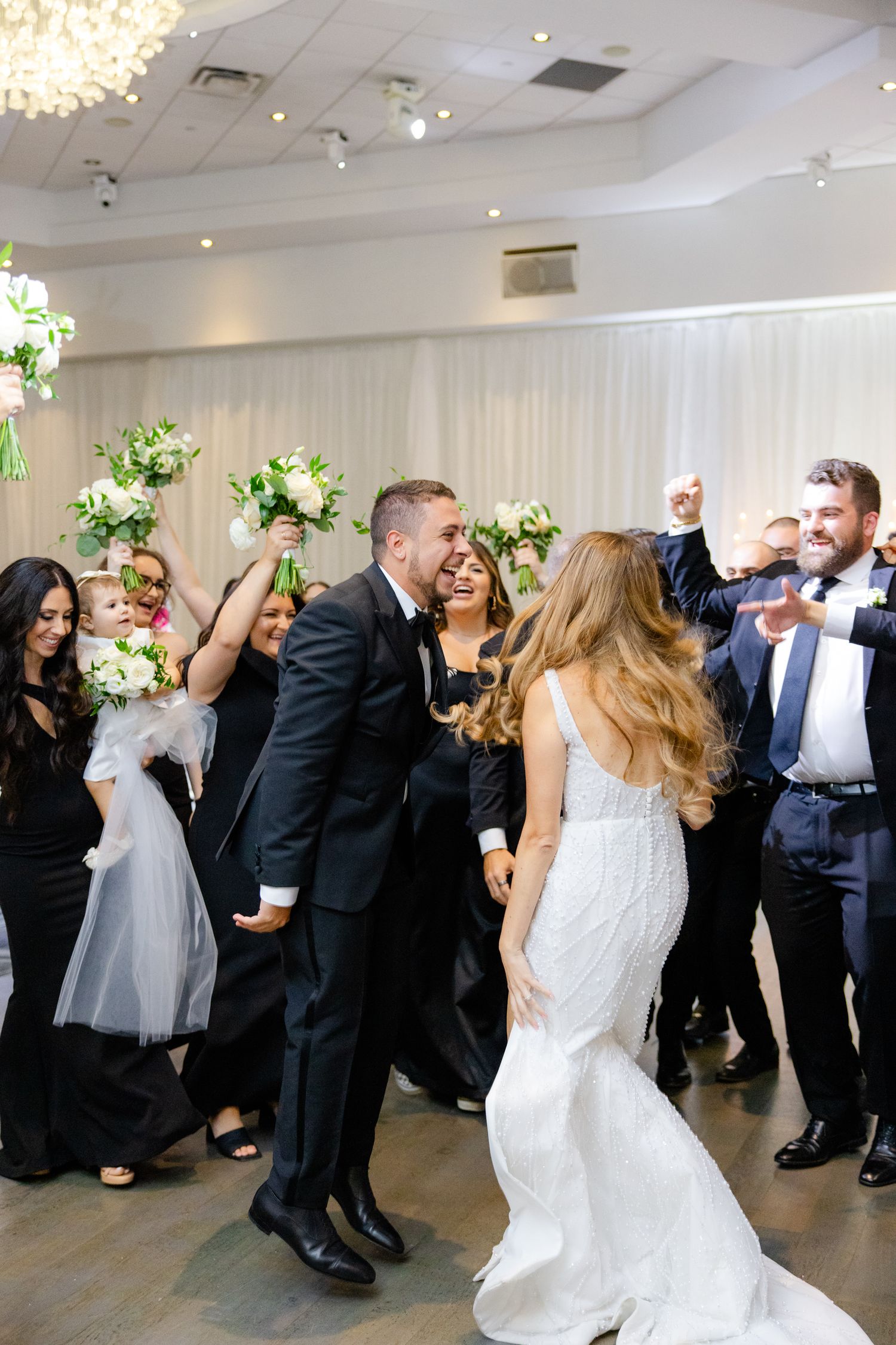 Wedding party dances and celebrates enthusiastically in modern reception hall with floral arrangements.