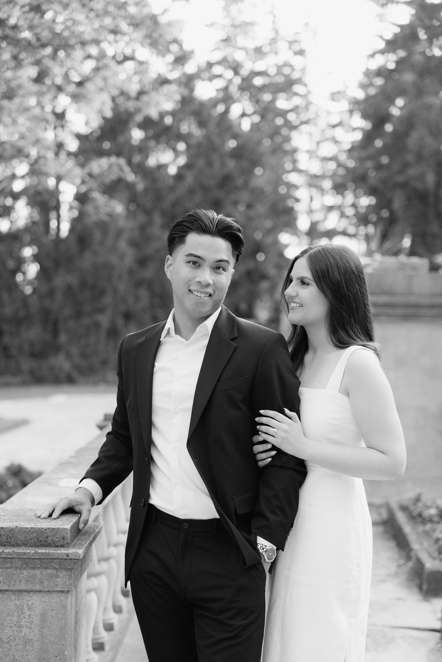 Black and white portrait of stylish couple posing by stone railing in garden setting.