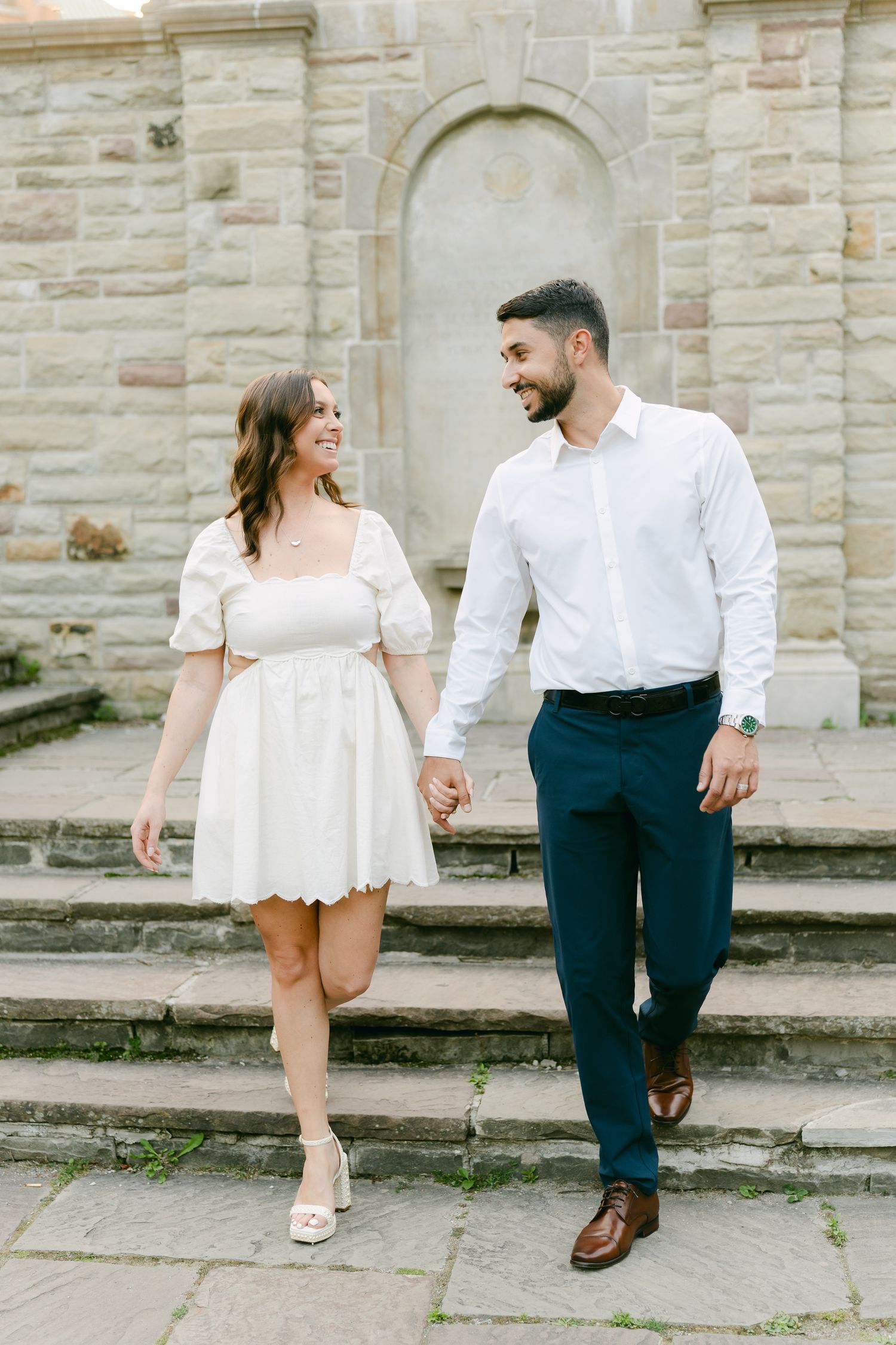 A romantic couple walks down stone steps while holding hands in coordinated formal attire.