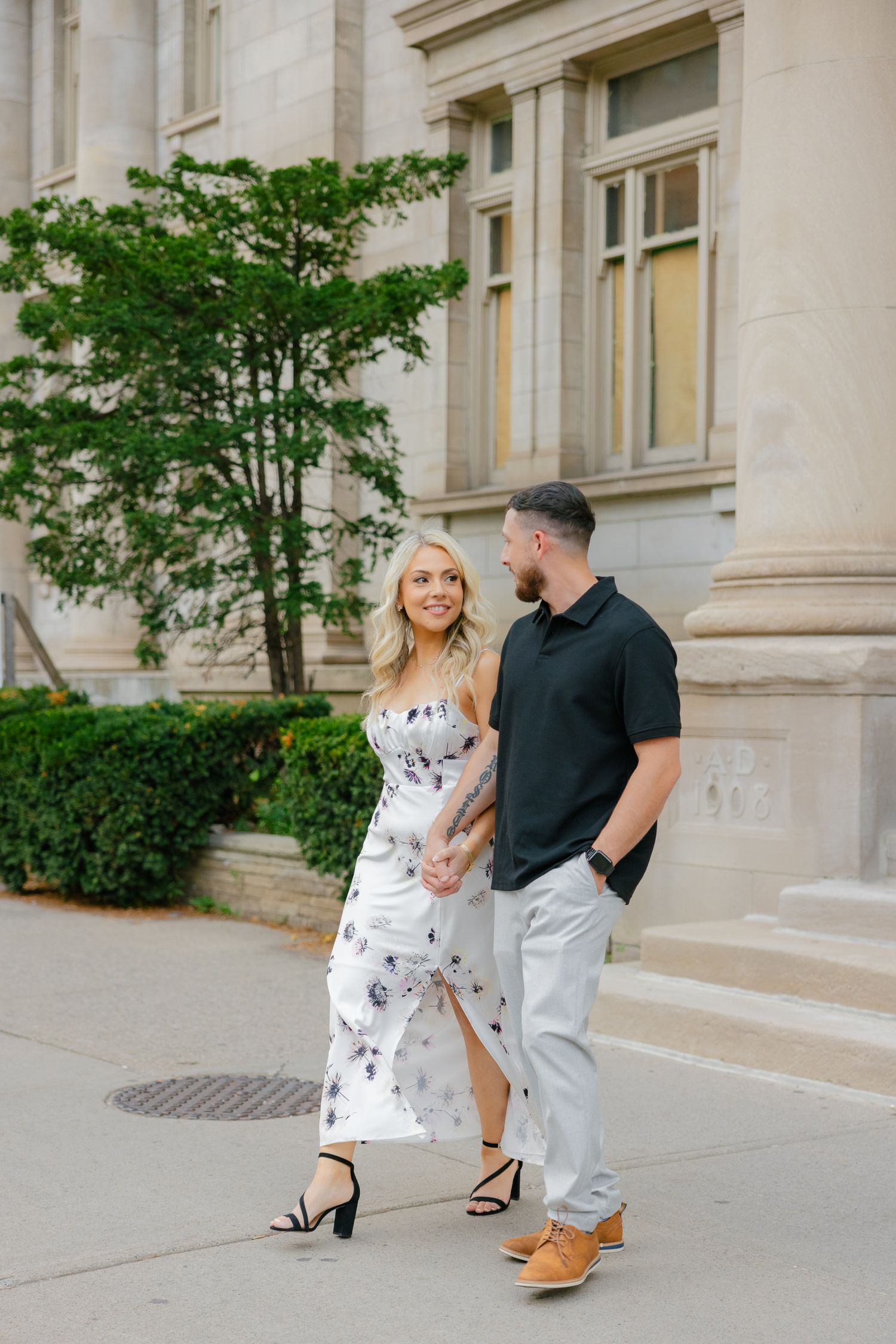 A couple strolls together beside a historic building with limestone columns and green trees.