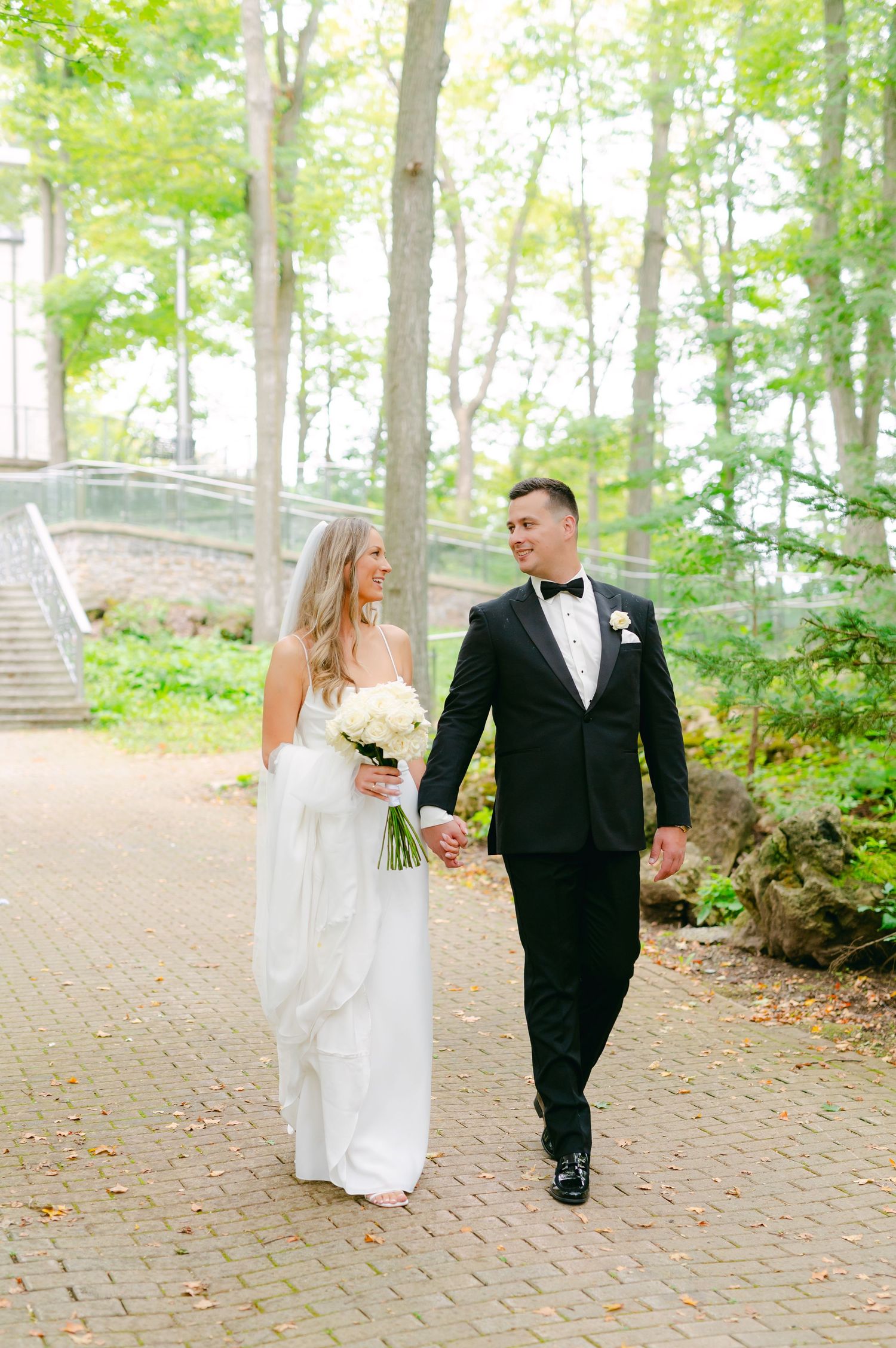 Newlyweds walk together on a brick path through a wooded area.