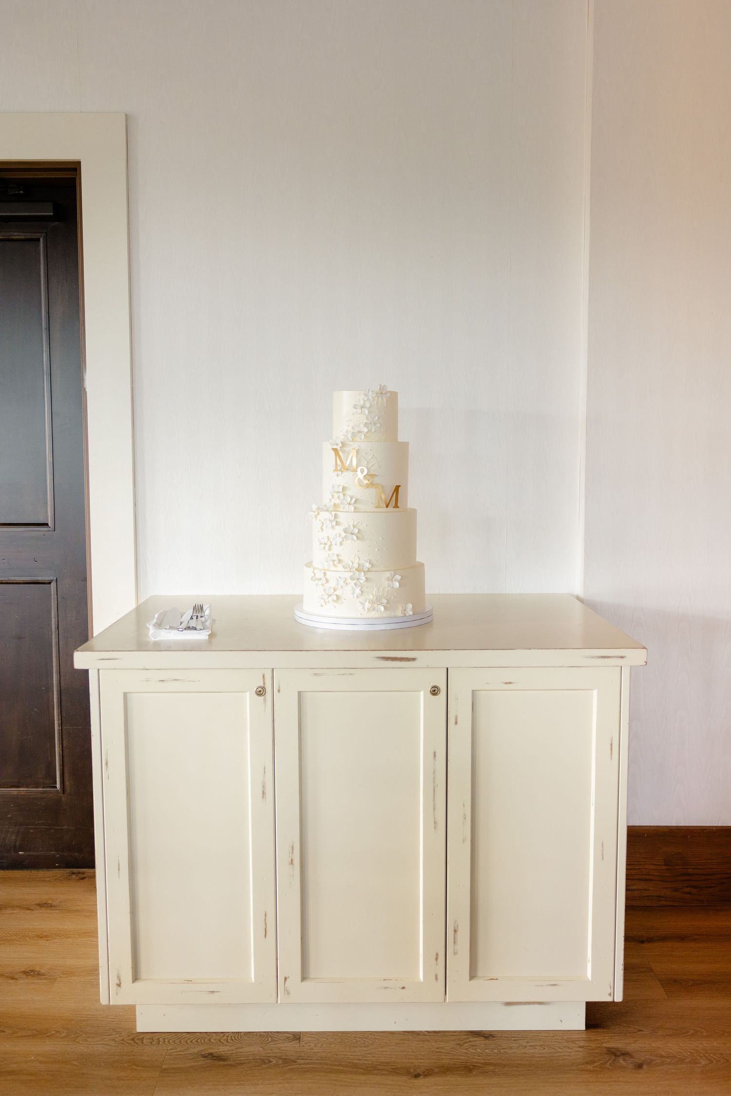 Elegant white wedding cake displayed on vintage white cabinet against neutral wall.