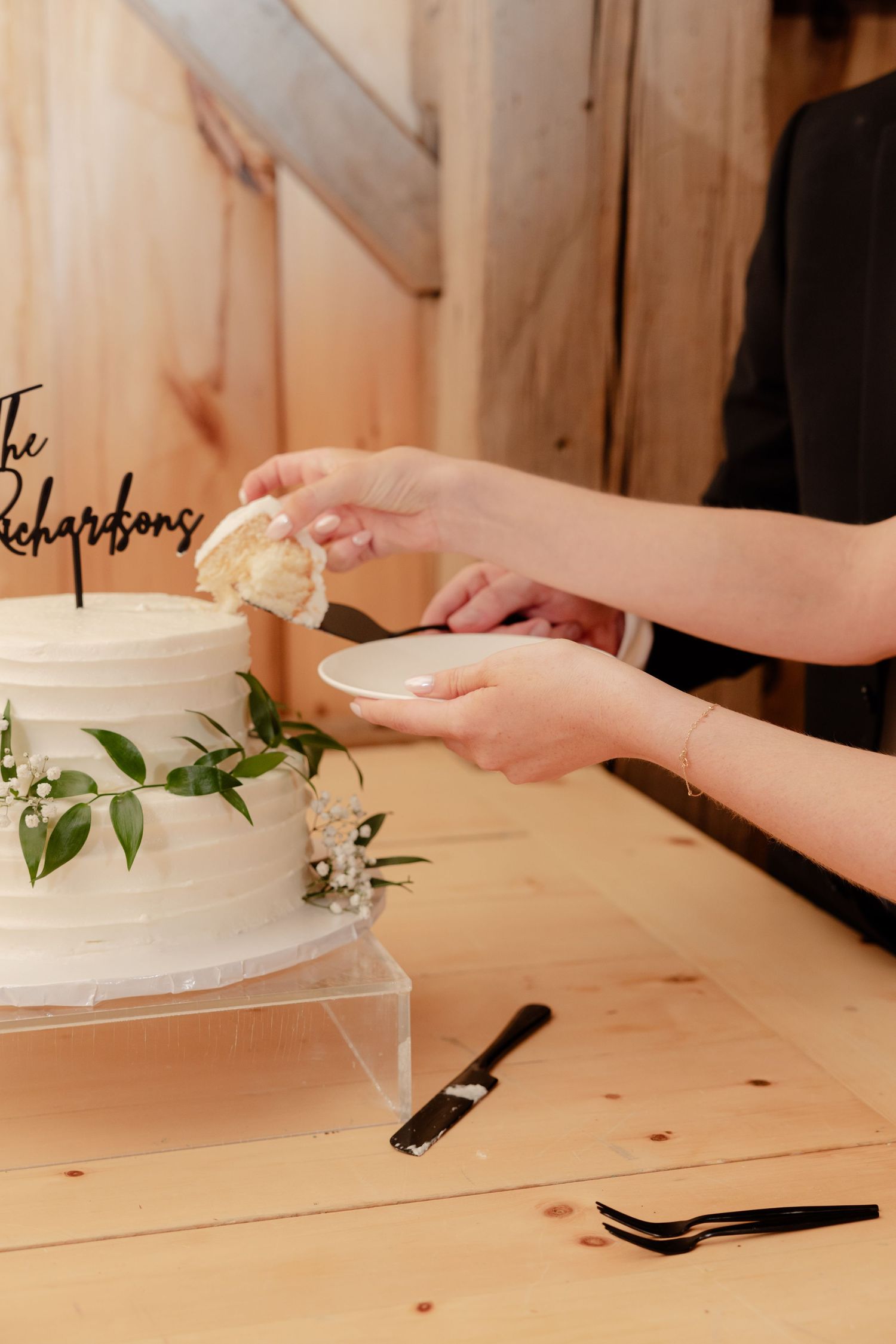 A pair of hands cutting into a white wedding cake decorated with greenery and a topper on a wooden table.