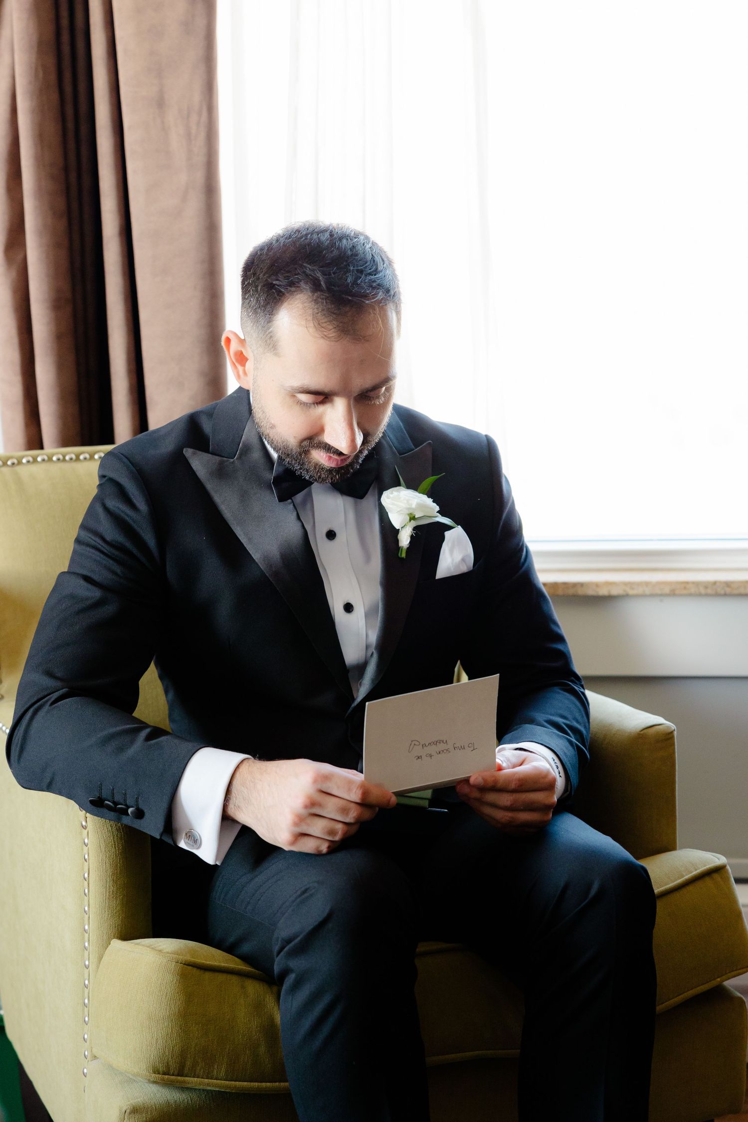 A person in a formal tuxedo sits in a green chair reading a note before a wedding.