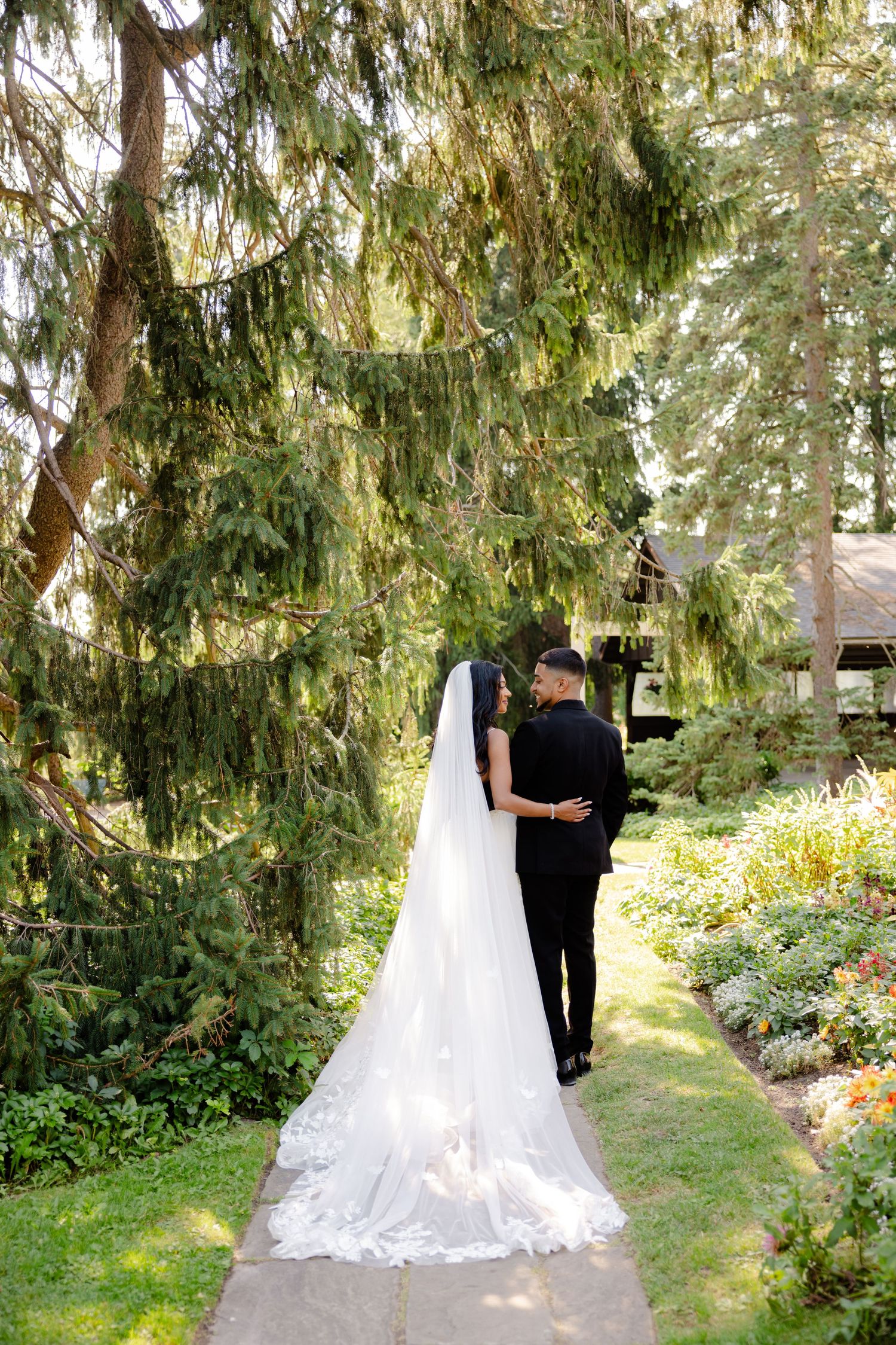 A couple shares an intimate moment on a garden path surrounded by towering evergreens.