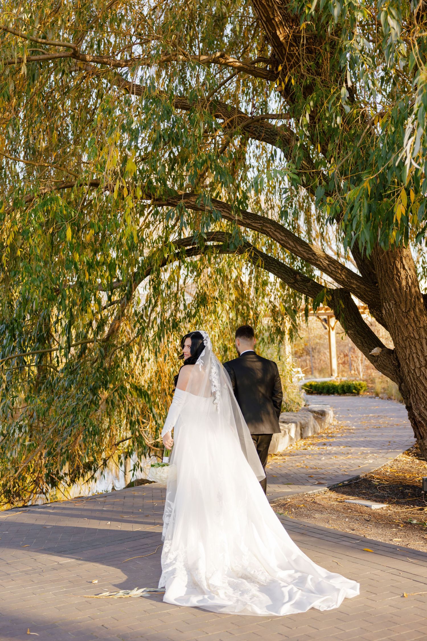A couple walks along a path under drooping willow branches on their wedding day.