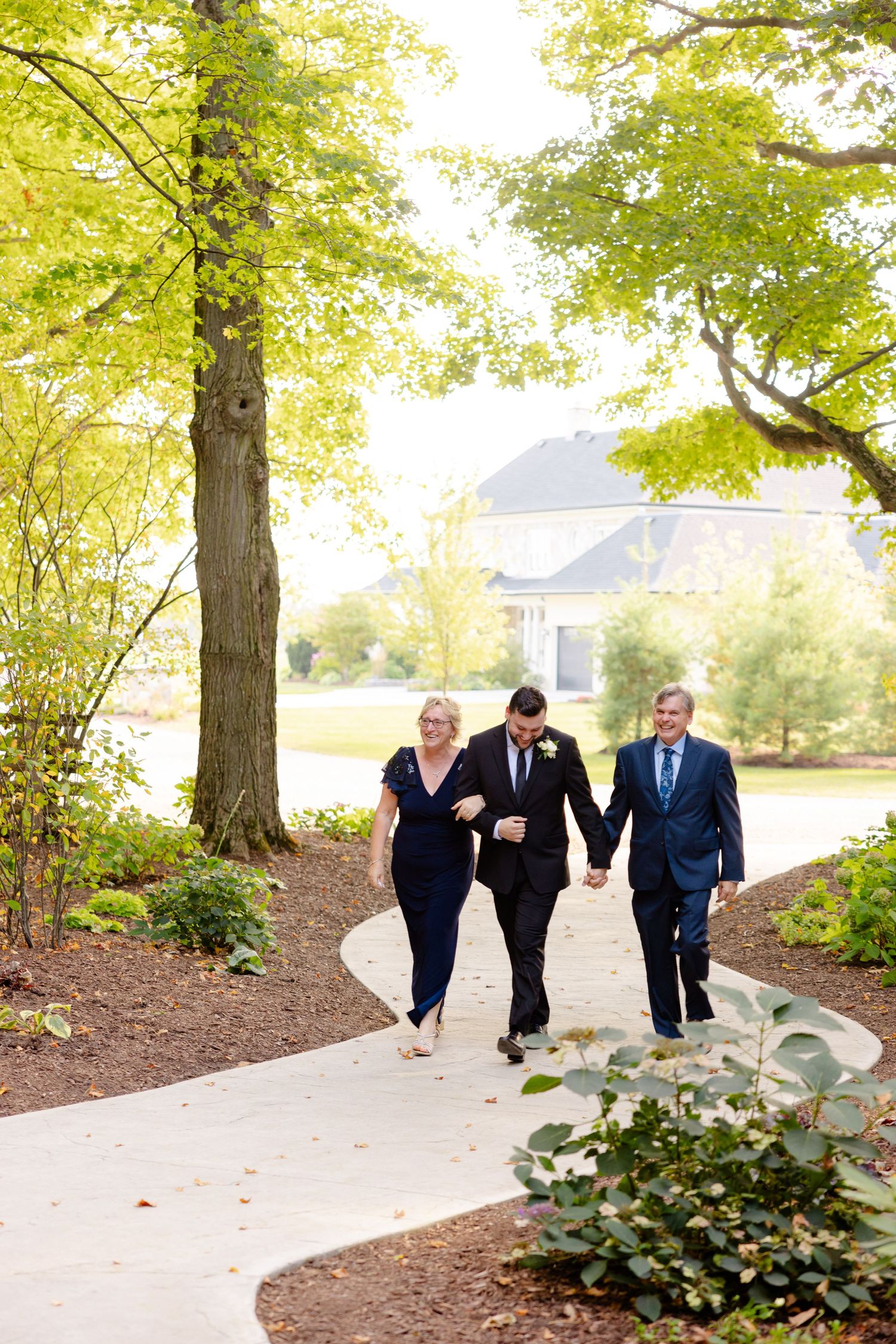 Wedding party members walk along a garden path with trees and landscaping in the background.