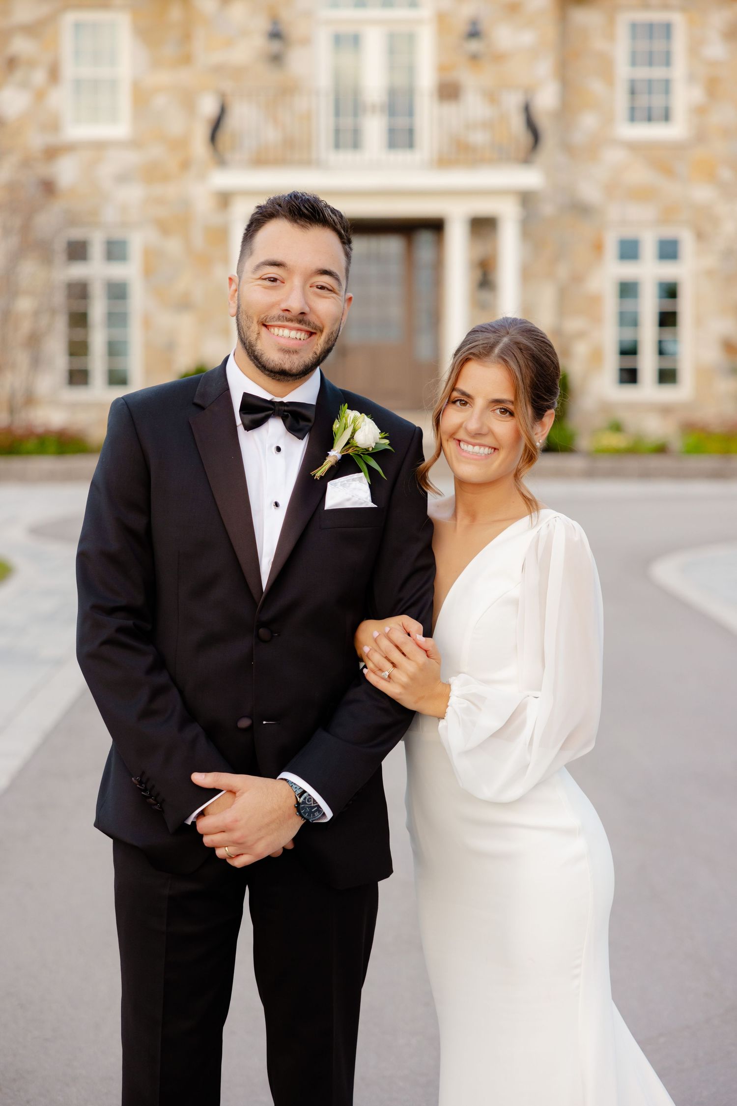 A happy couple in formal wedding attire poses in front of a grand stone house.