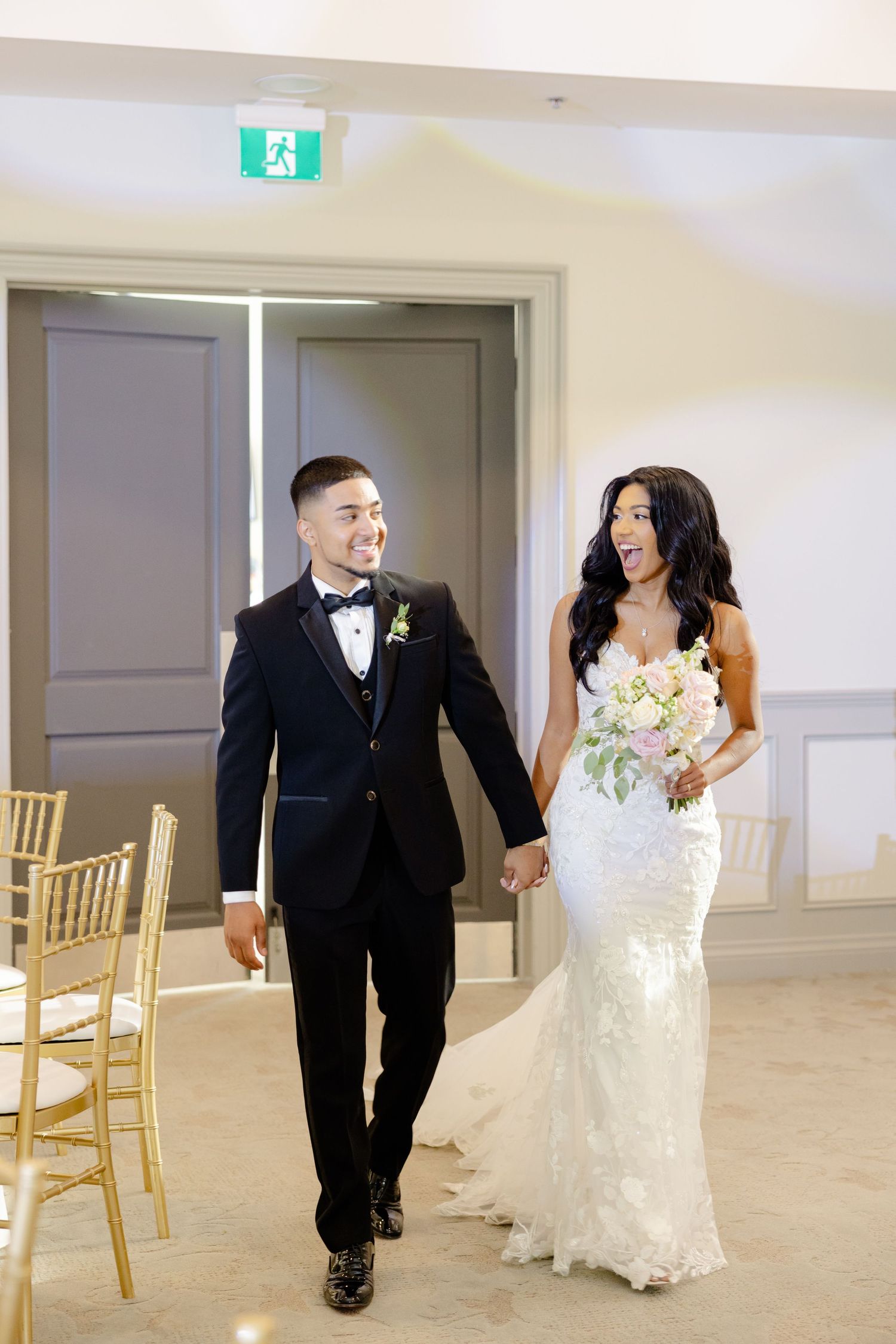 Elegant wedding couple walking together in a bright indoor venue with gold chairs.