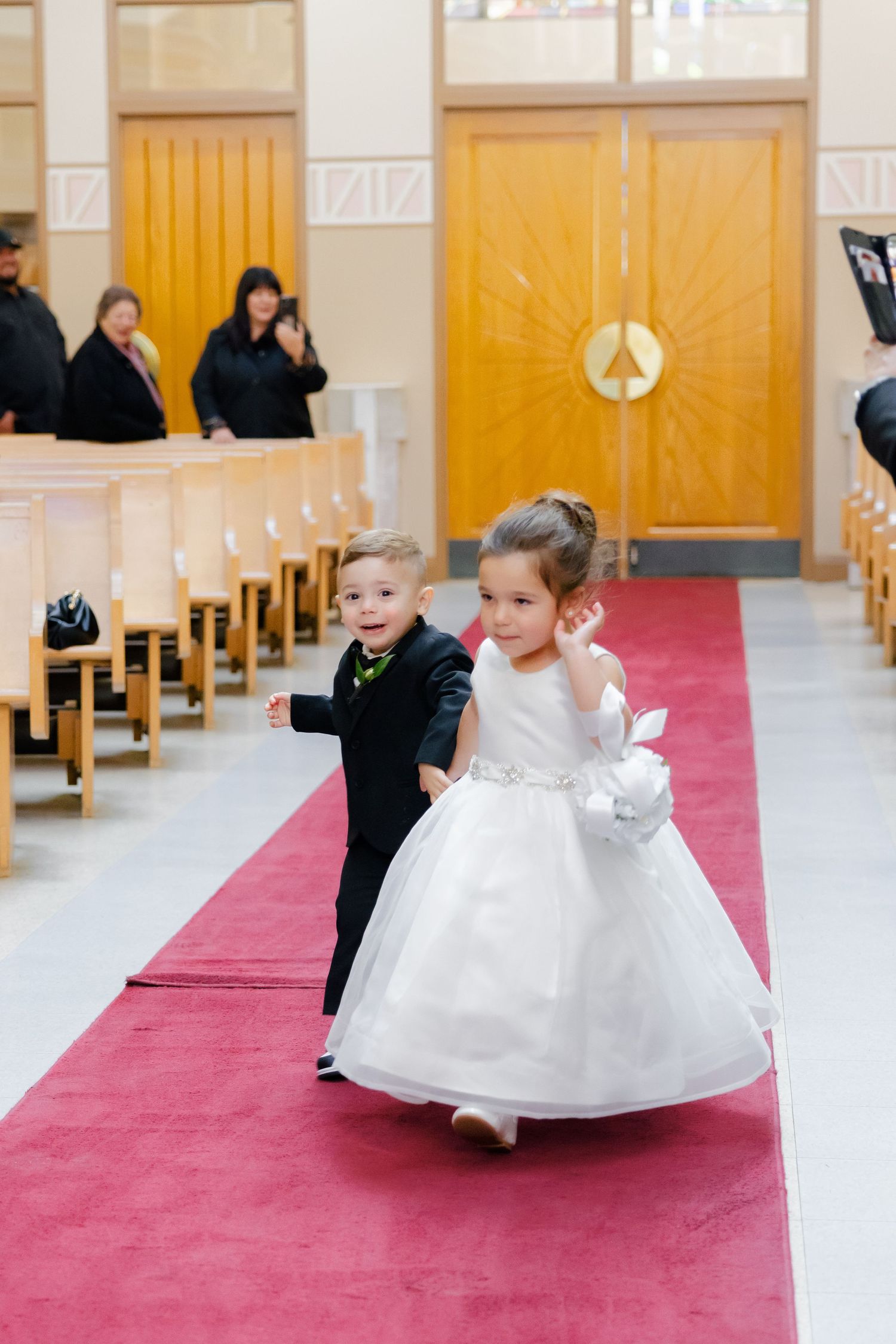 A young ring bearer and flower girl in formal attire walk down a red-carpeted church aisle.
