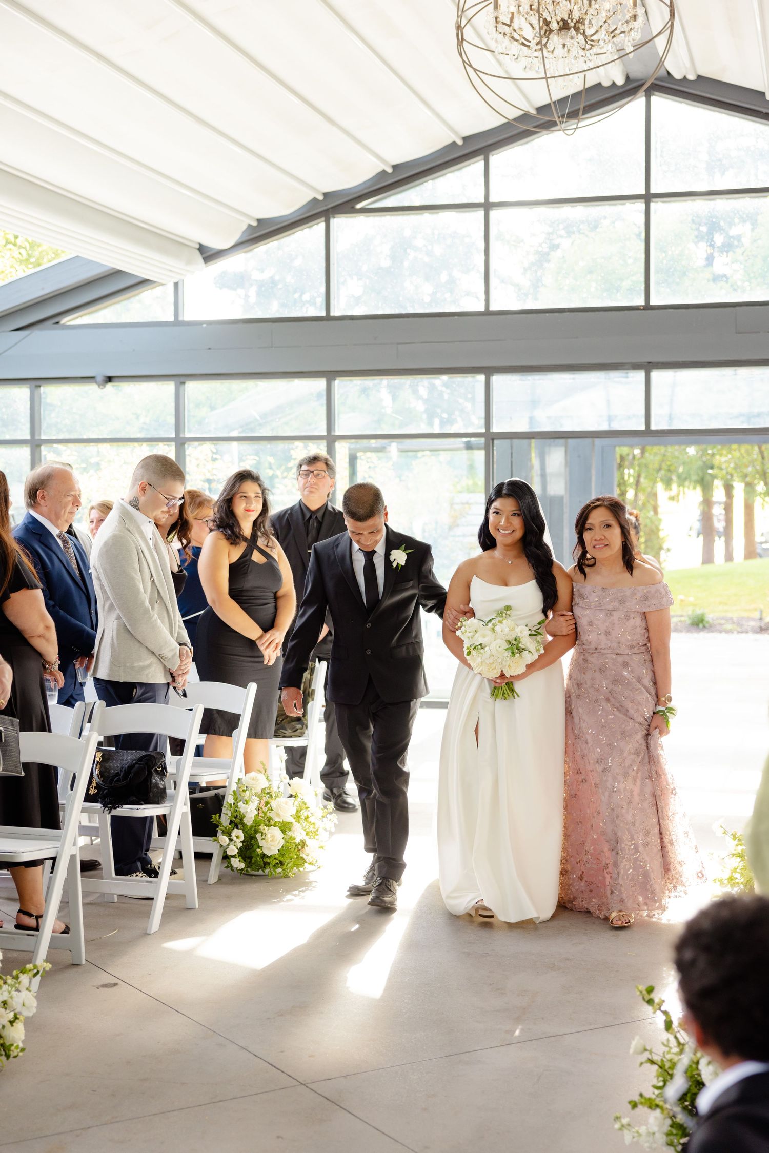 A wedding party walks down the aisle in a light-filled venue decorated with white chairs and flowers.
