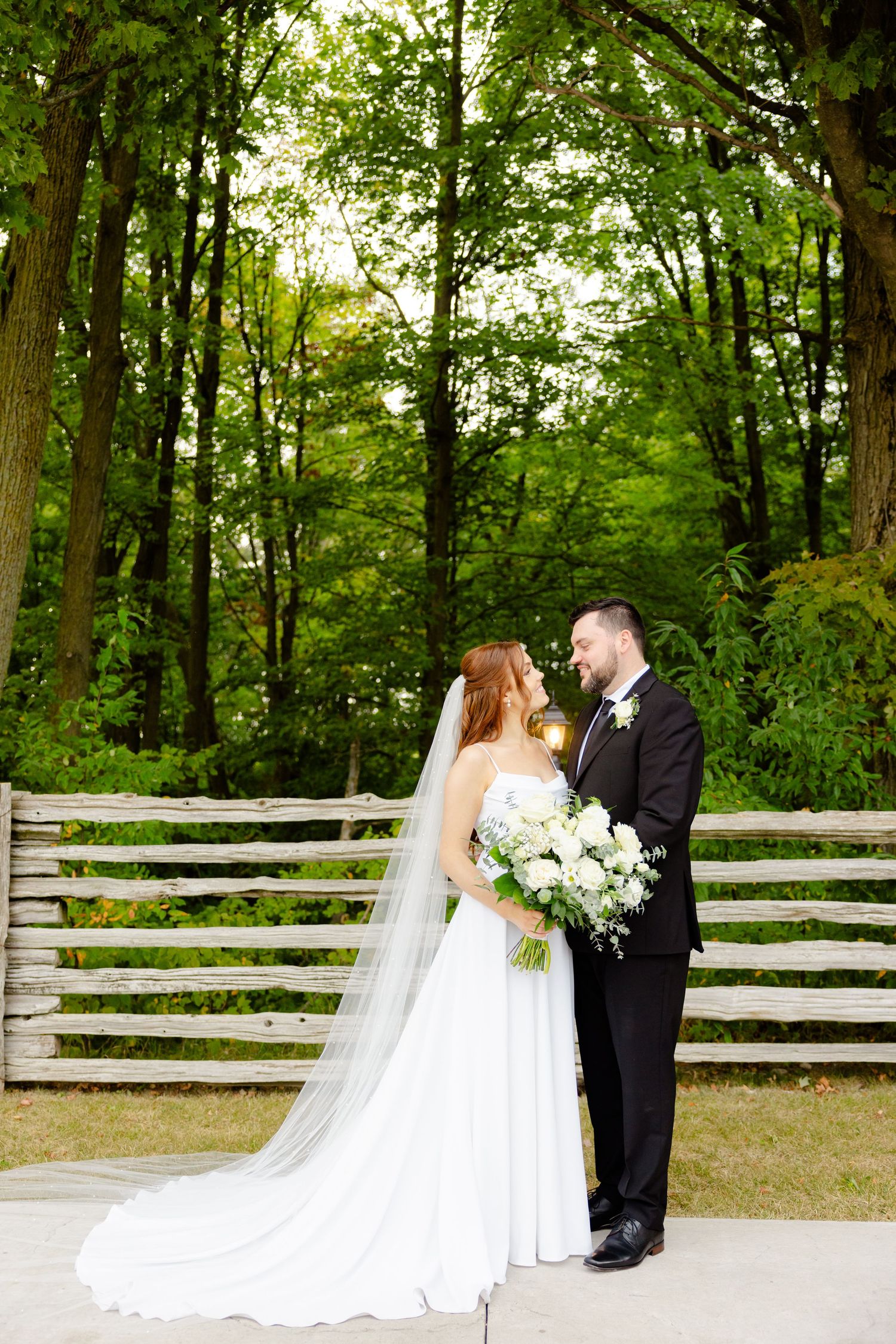 A bride and groom pose together outdoors with a beautiful white floral bouquet against a wooded backdrop.