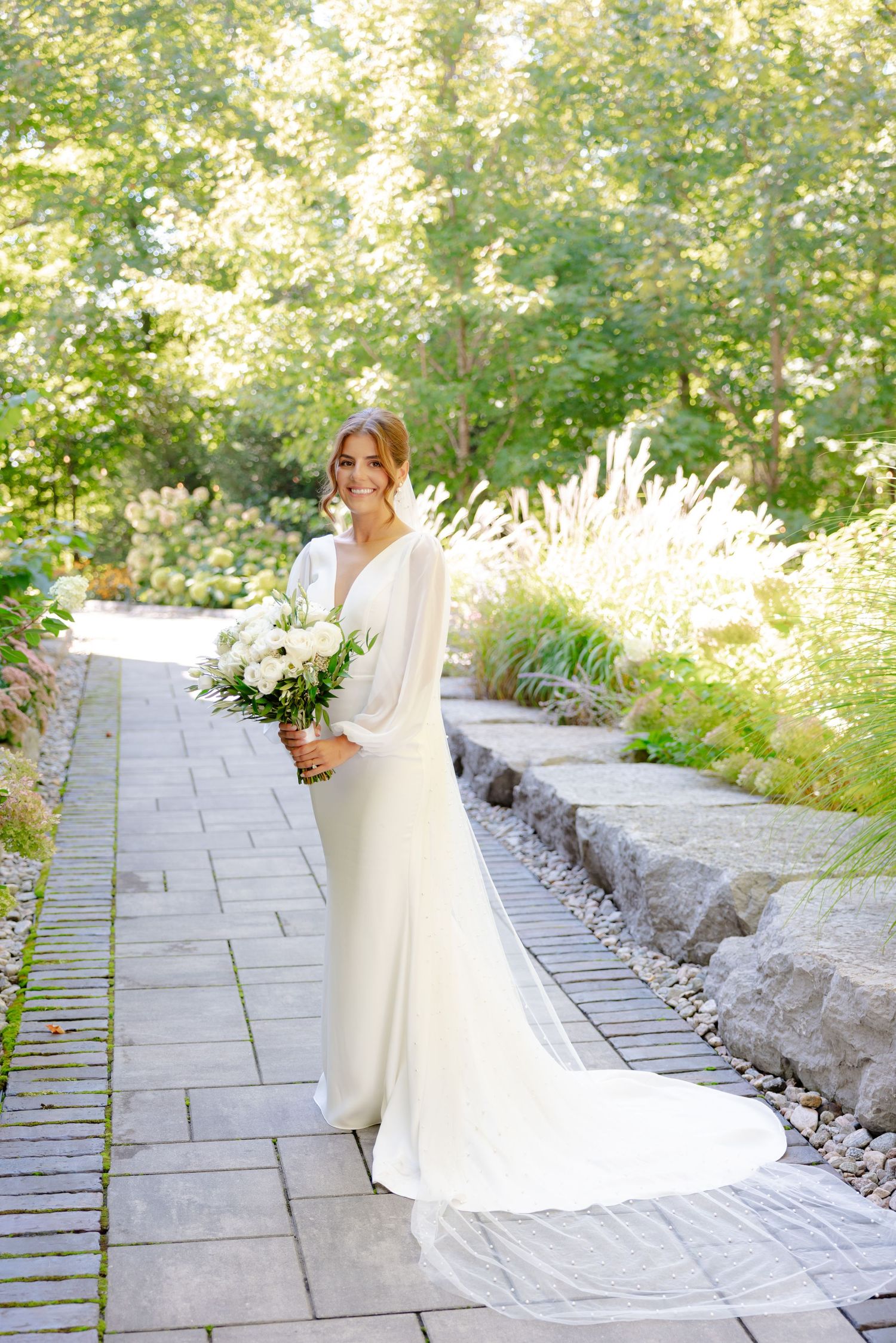 Elegant bride in long-sleeved white dress holding white bouquet on stone garden path.