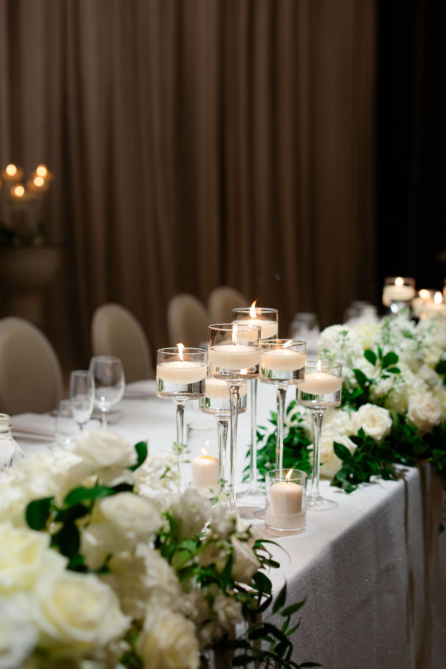 Elegant wedding table setting with floating candles and white rose centerpieces against a brown curtained backdrop.