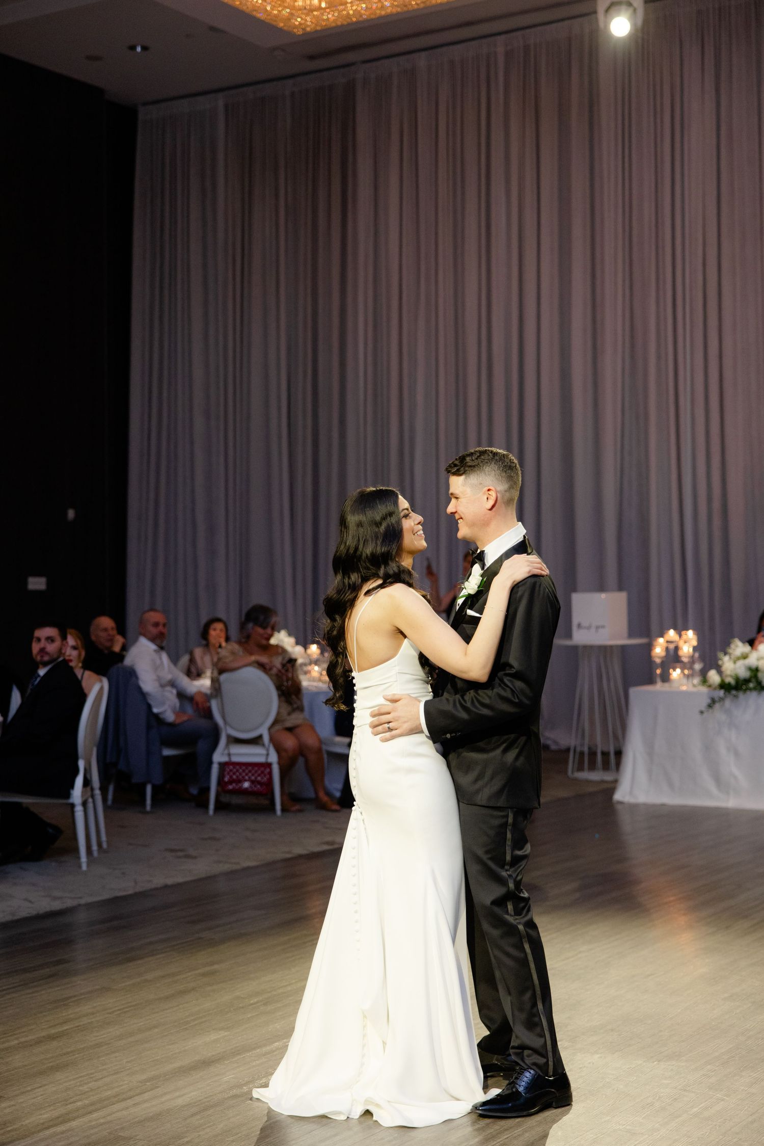 A couple shares their first dance as newlyweds in an elegant ballroom with floor-to-ceiling curtains.