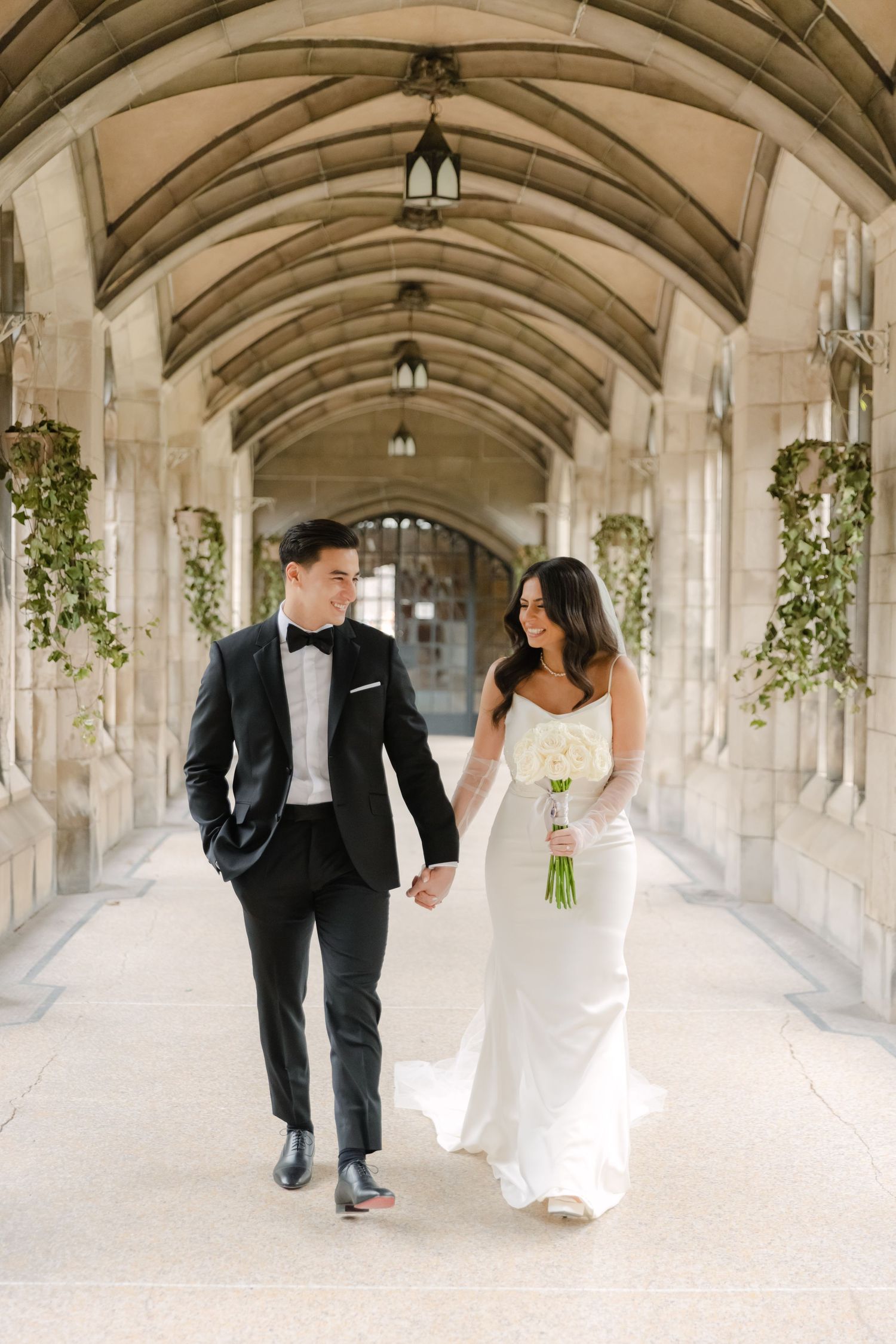 Newlyweds walk hand in hand through elegant stone archway with hanging greenery and lanterns.