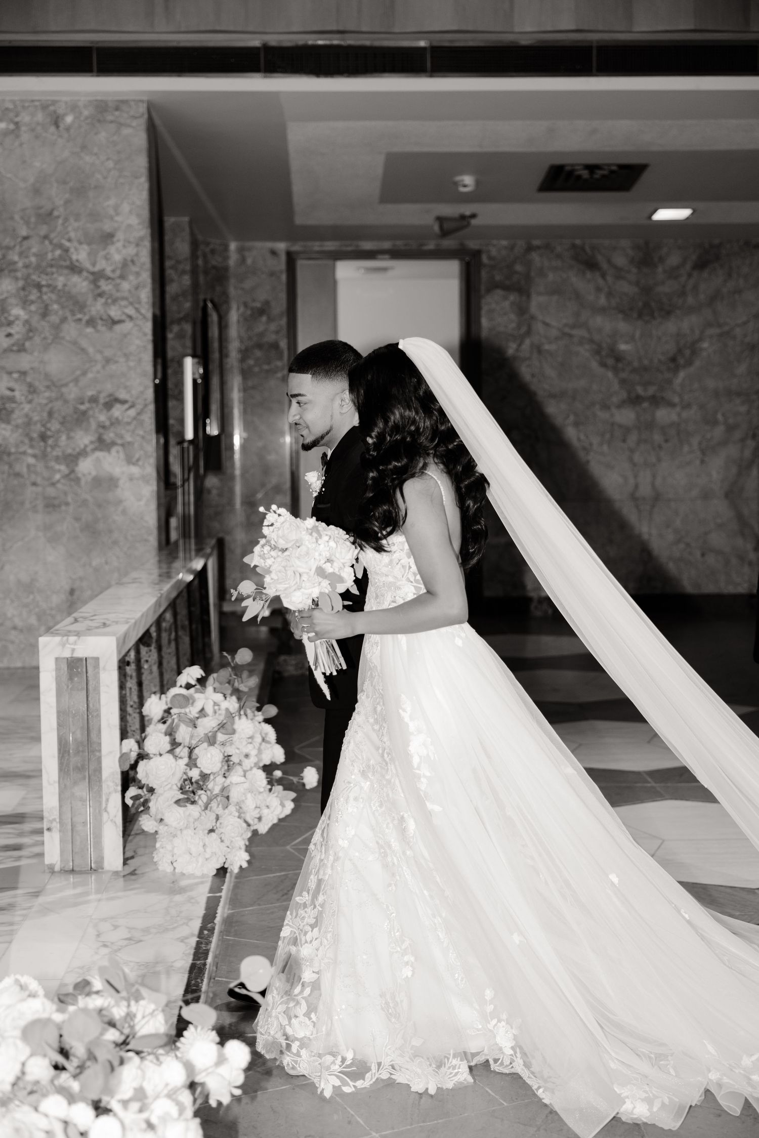 Elegant bride in lace gown with flowing cathedral veil holds white bouquet in marble hallway.