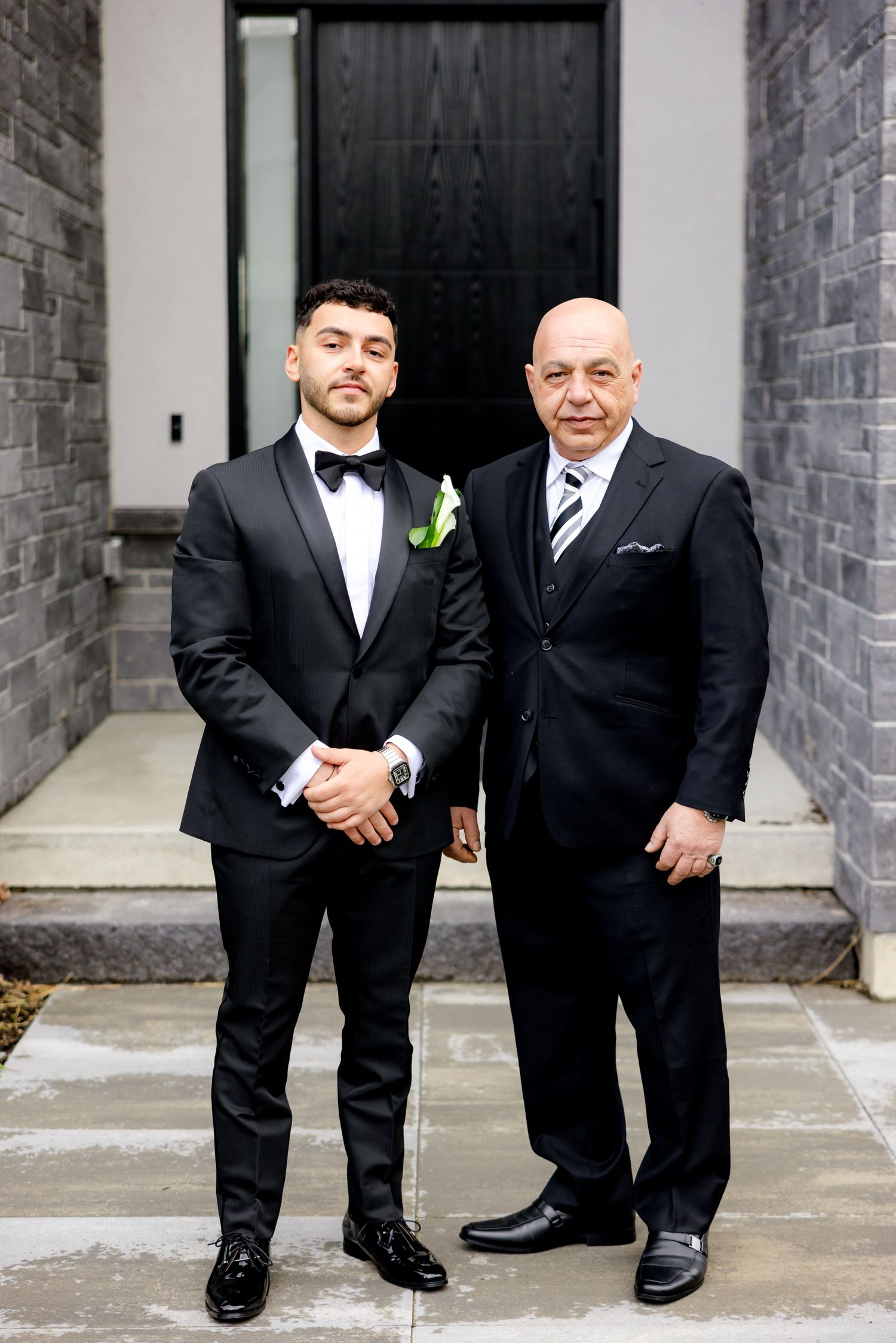 Formal portrait of two men in black suits standing in front of modern doorway.