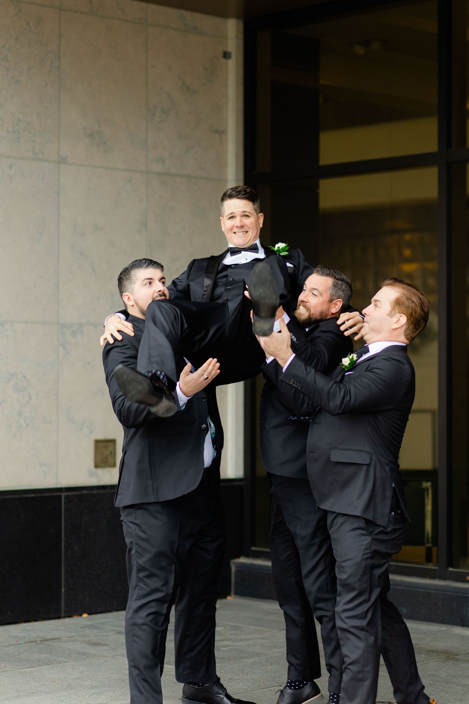 Four groomsmen in formal attire lift and hold up one of their party members while smiling and laughing.