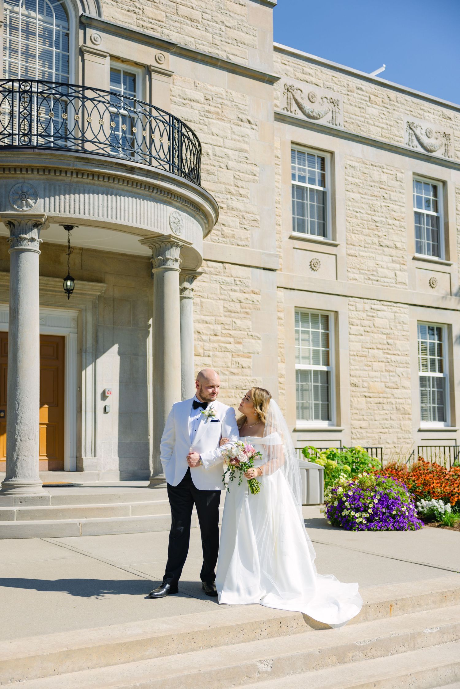 Wedding couple poses on steps of elegant stone building with curved balcony and colorful garden.