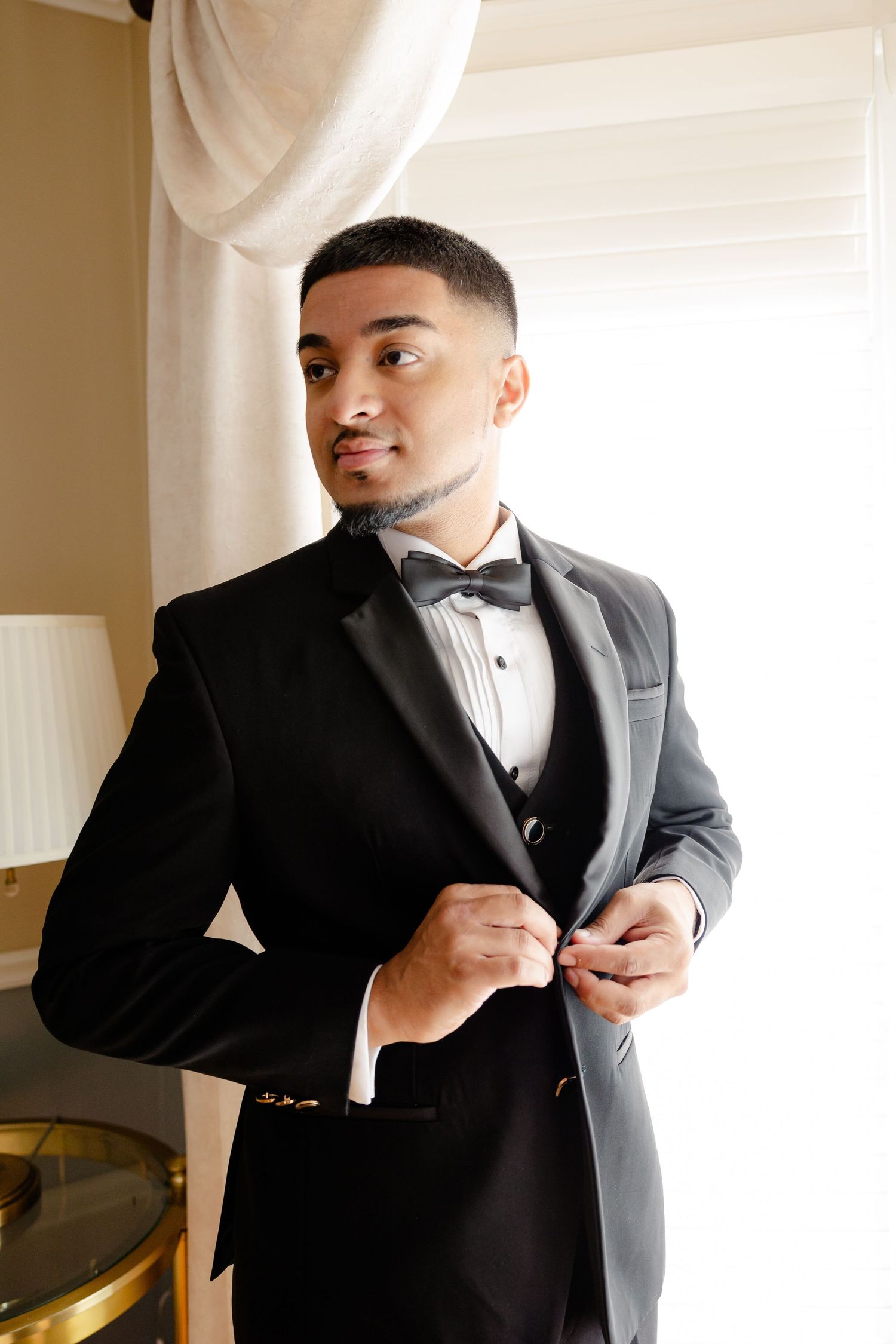 A person in a black tuxedo and bow tie adjusts their jacket in front of soft window light.