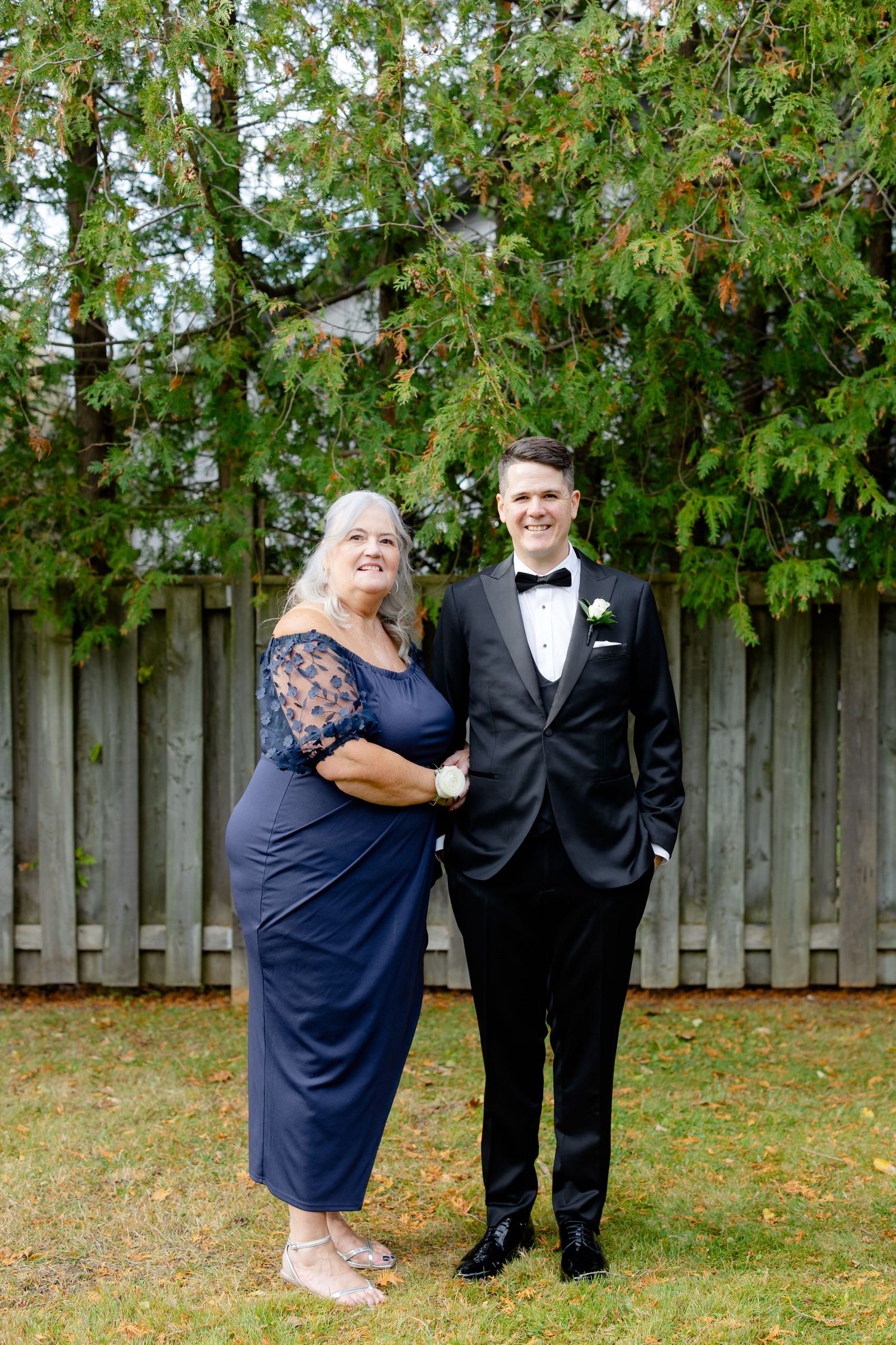 A formal portrait of two people in elegant attire posing together against a wooden fence and green foliage.