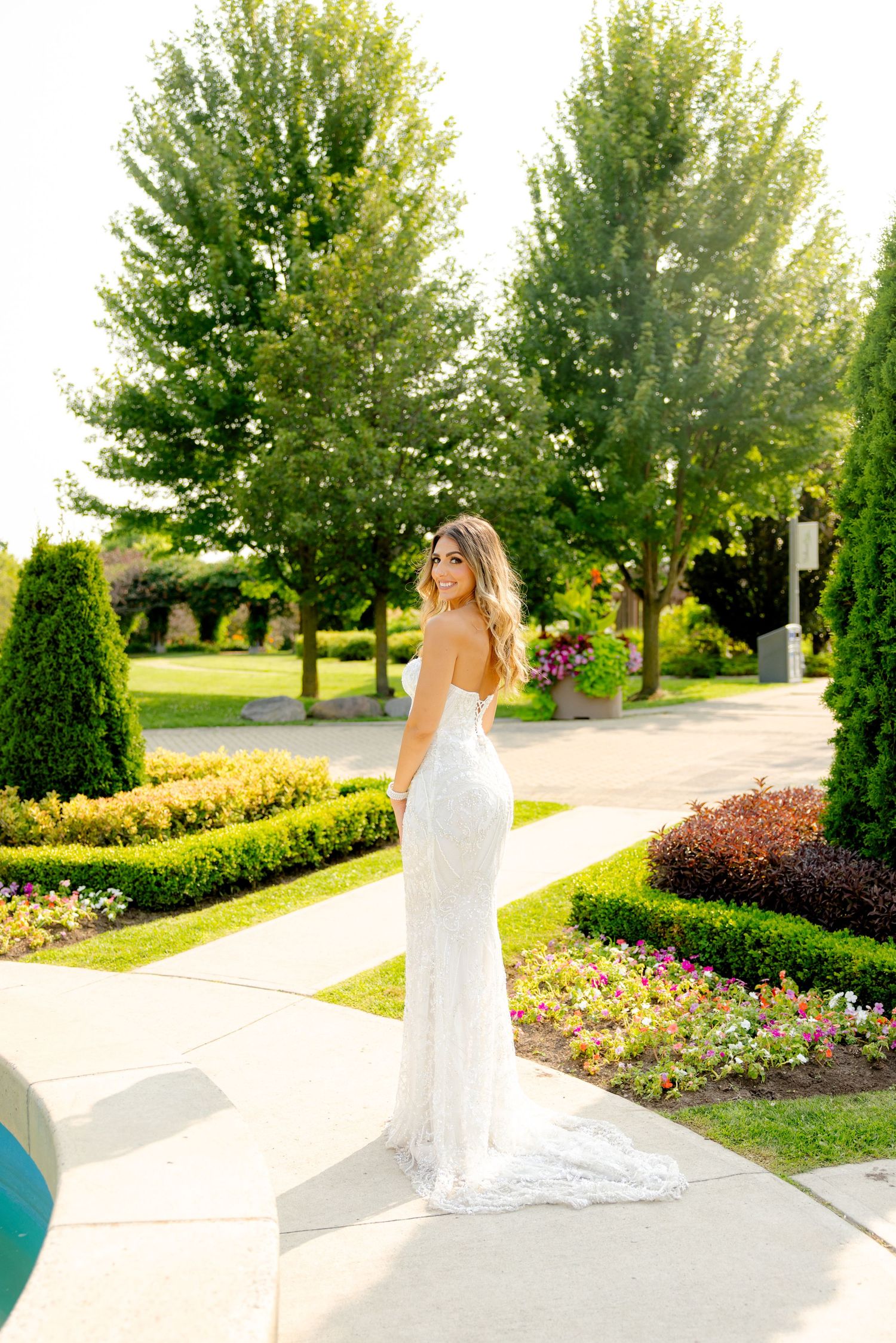 Elegant bride in white gown stands on pathway surrounded by landscaped garden and trees.