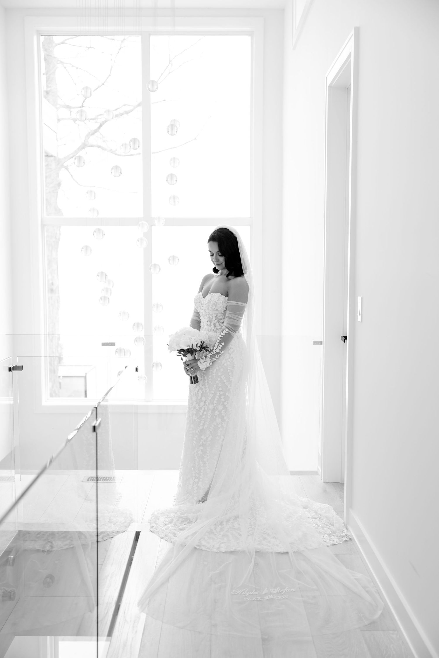 A black and white bridal portrait showing an elegant lace gown with long train in a sunlit hallway.