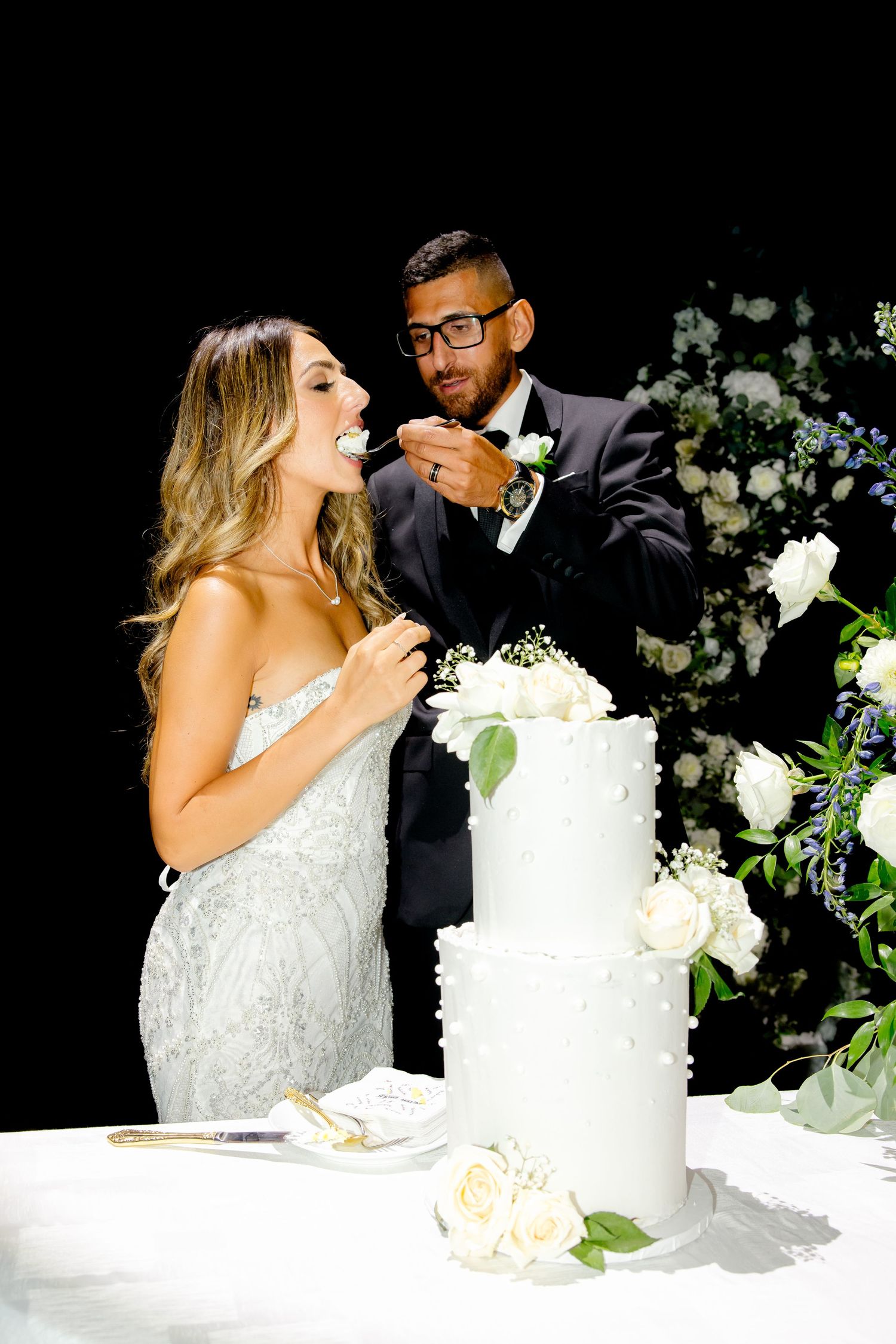Candid moment of newlyweds sharing wedding cake with white floral decorations.