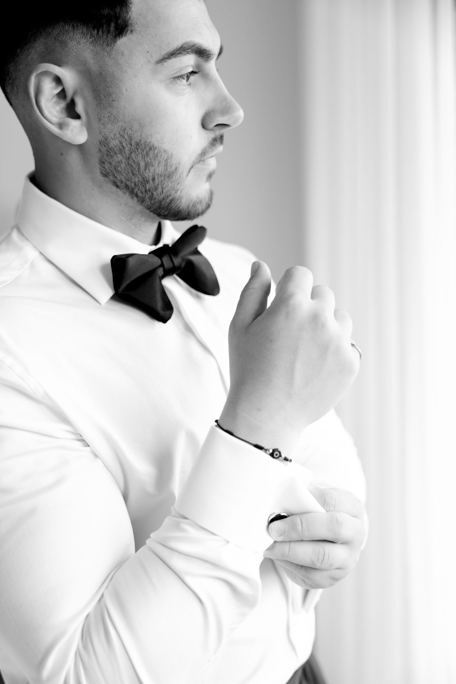 Profile view of person in formal attire adjusting bow tie near window in black and white.