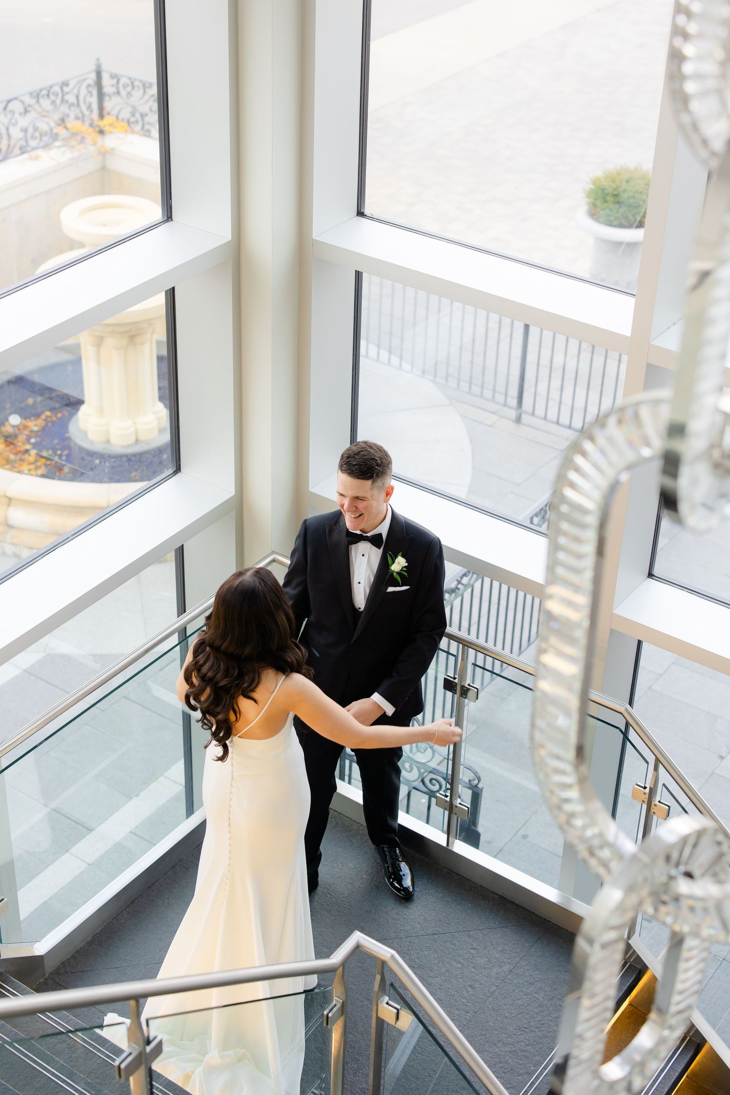 A couple shares an intimate wedding moment on a modern staircase with large windows and crystal chandelier details.