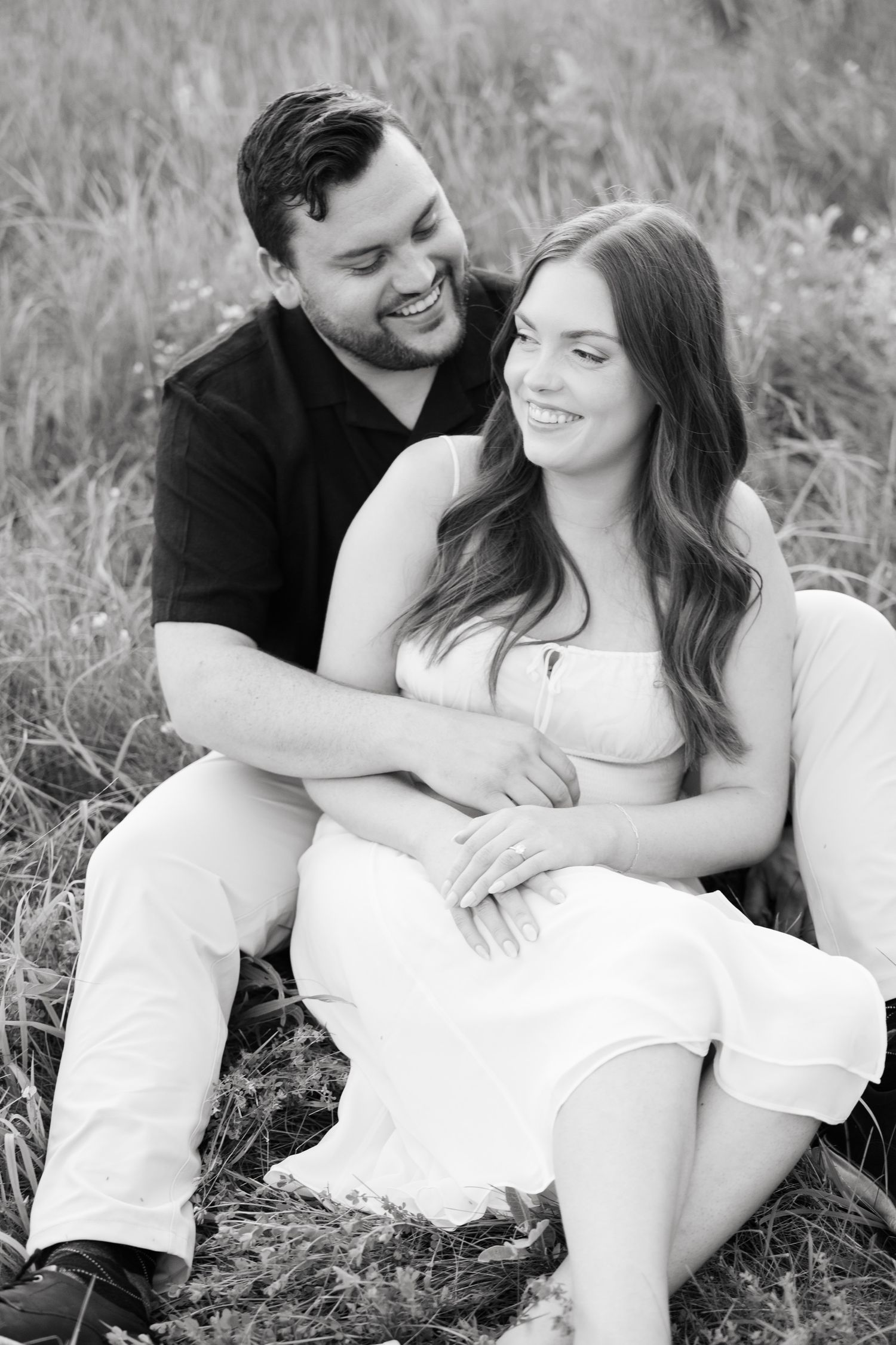 Romantic couple sits together in a grassy field sharing a tender moment in black and white photography.