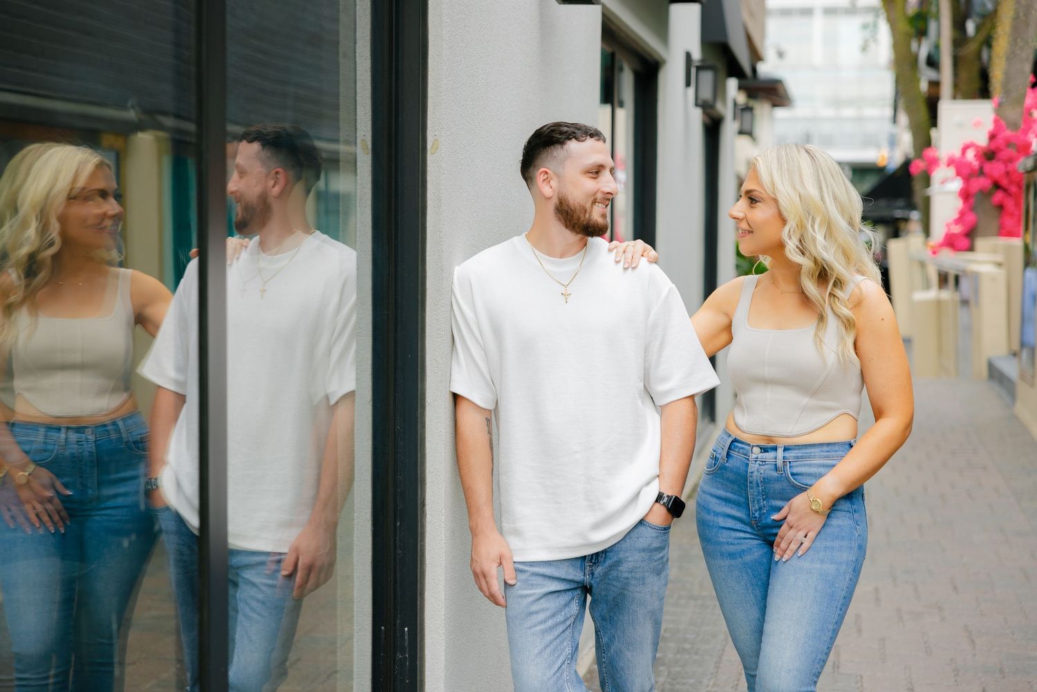 A couple in casual white shirts and jeans stroll together along a storefront window on an urban sidewalk.