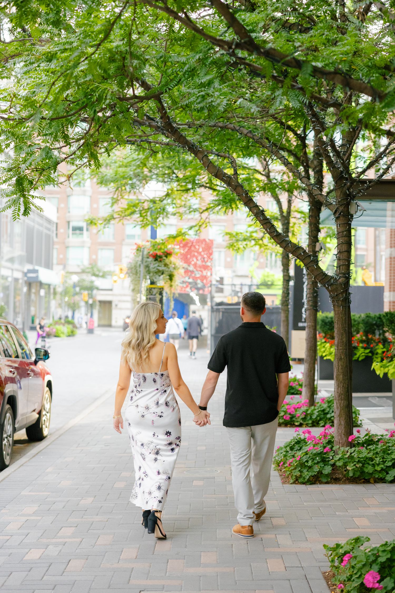 Couple walking hand in hand down a tree-lined urban sidewalk on a sunny day.