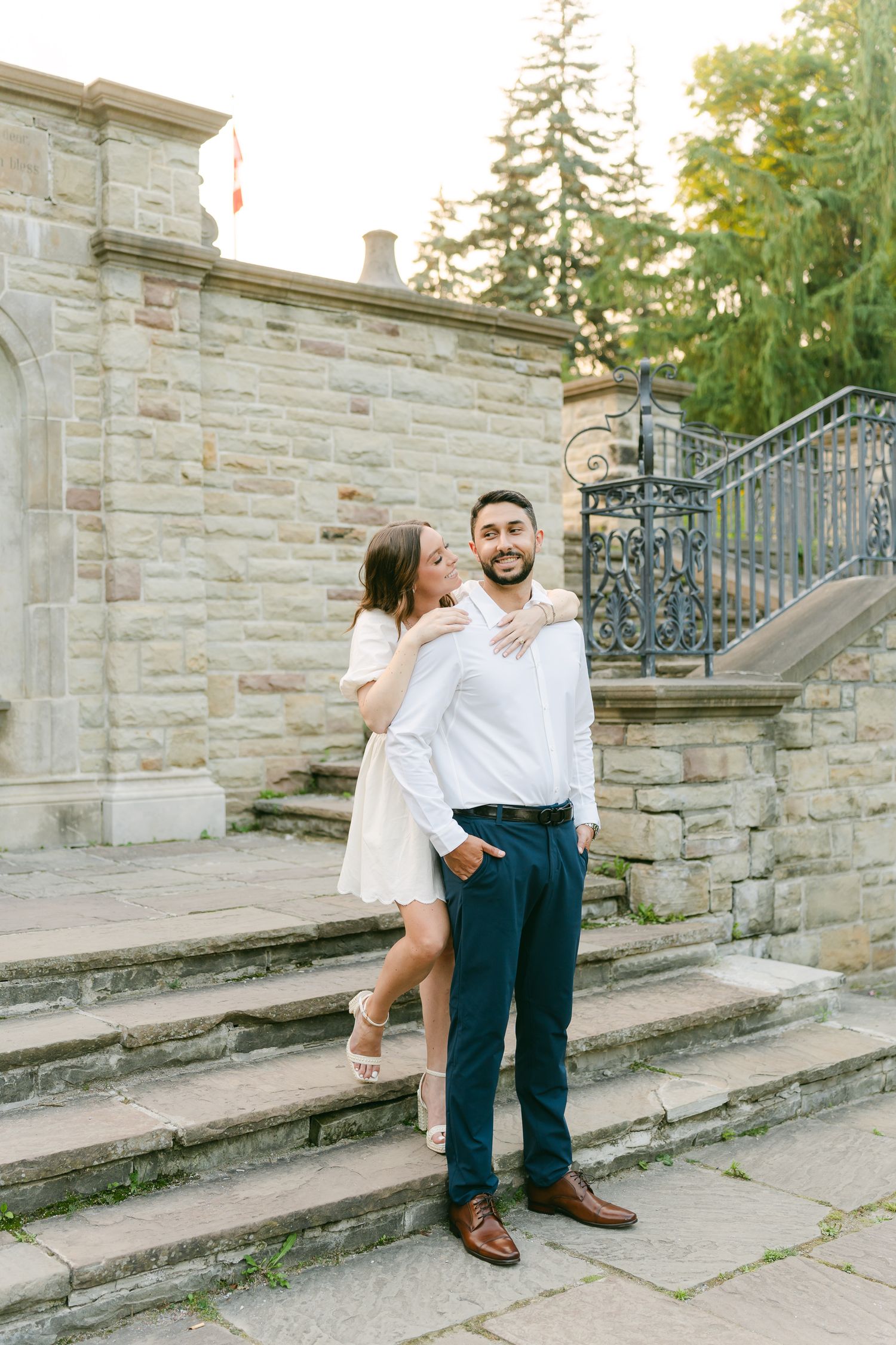 A playful couple poses on stone steps with ornate iron railings in matching white outfits and navy pants.
