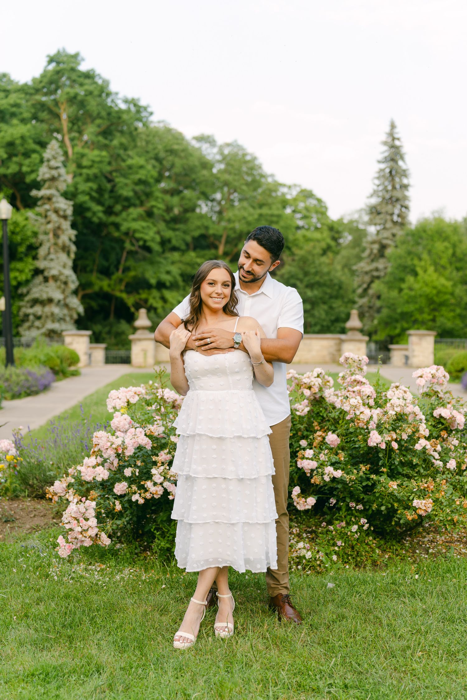 A couple poses together in a manicured garden with stone pillars and blooming roses.