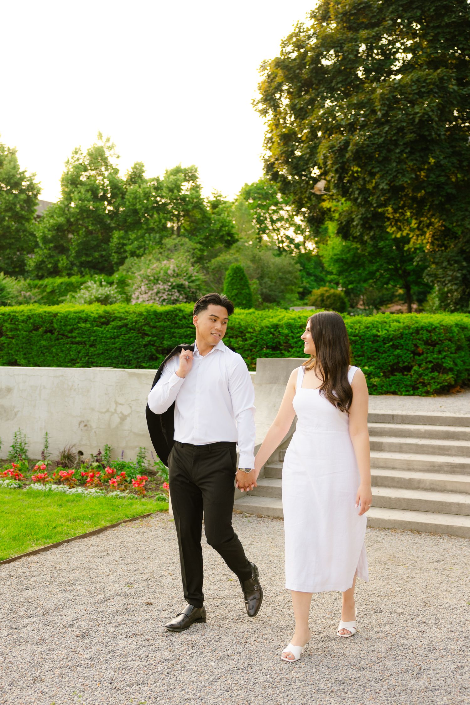 Couple strolls hand in hand near stone steps with flowering gardens and manicured hedges.