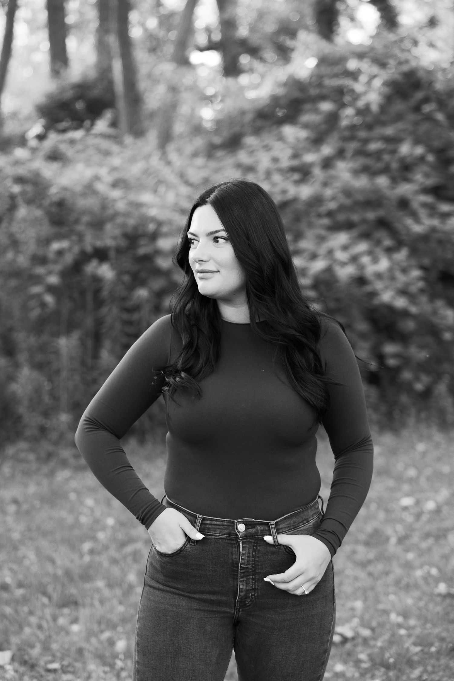 Woman in black long sleeve top and jeans poses confidently in black and white forest portrait.