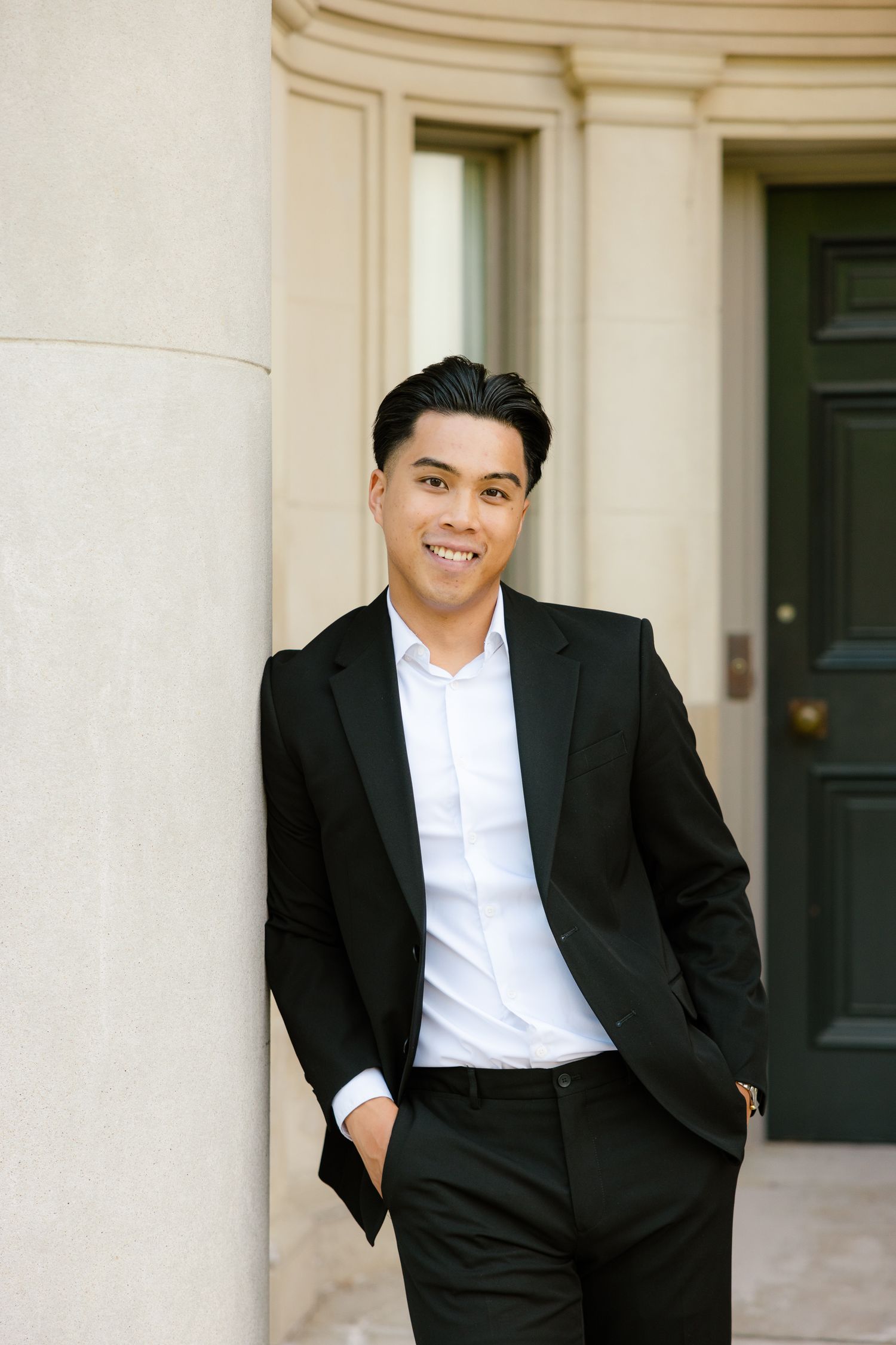 Portrait of well-dressed individual in black suit and white shirt standing by classical column and doorway.