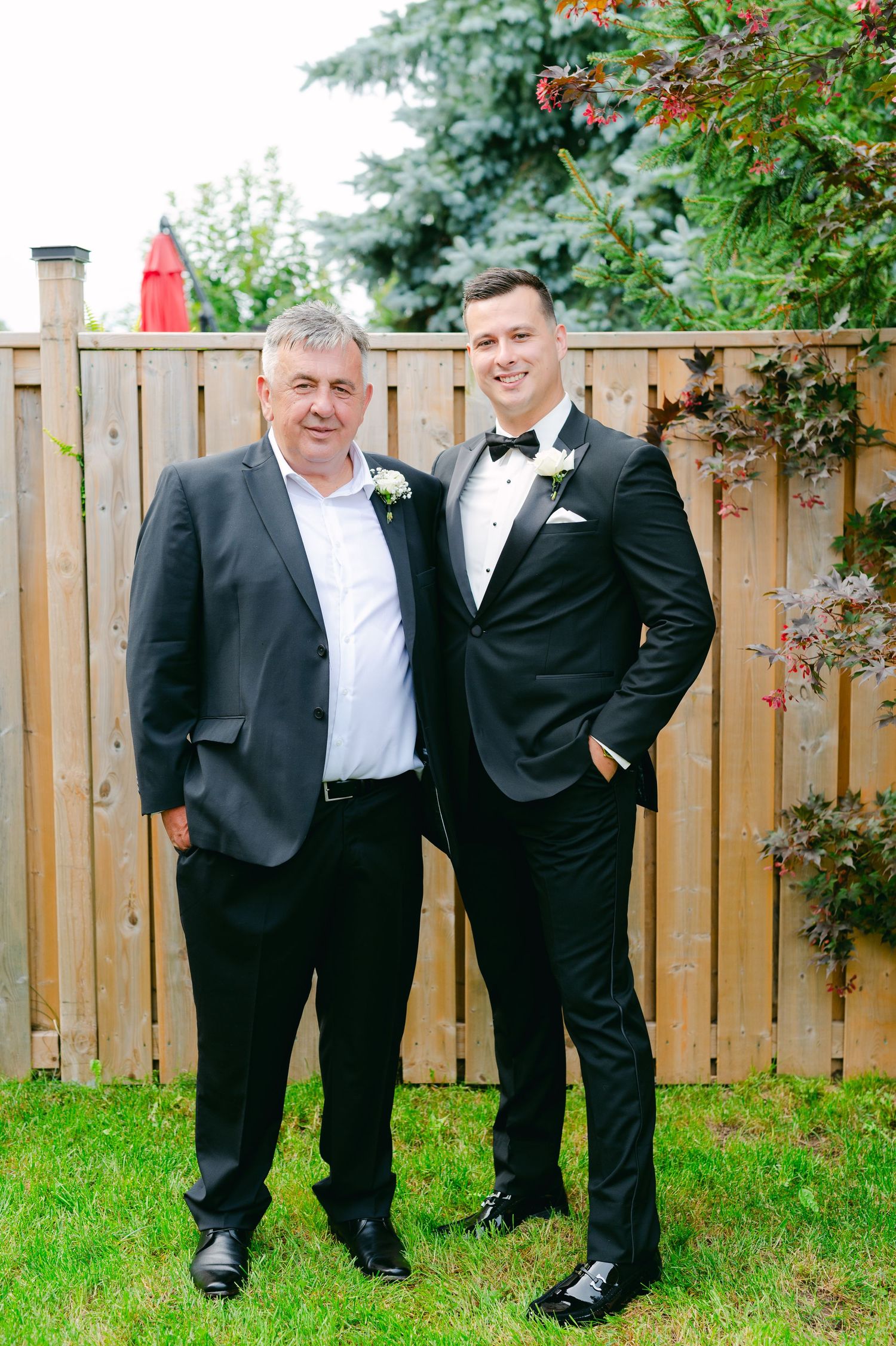 Two people in formal suits stand together in front of a wooden fence at an outdoor event.