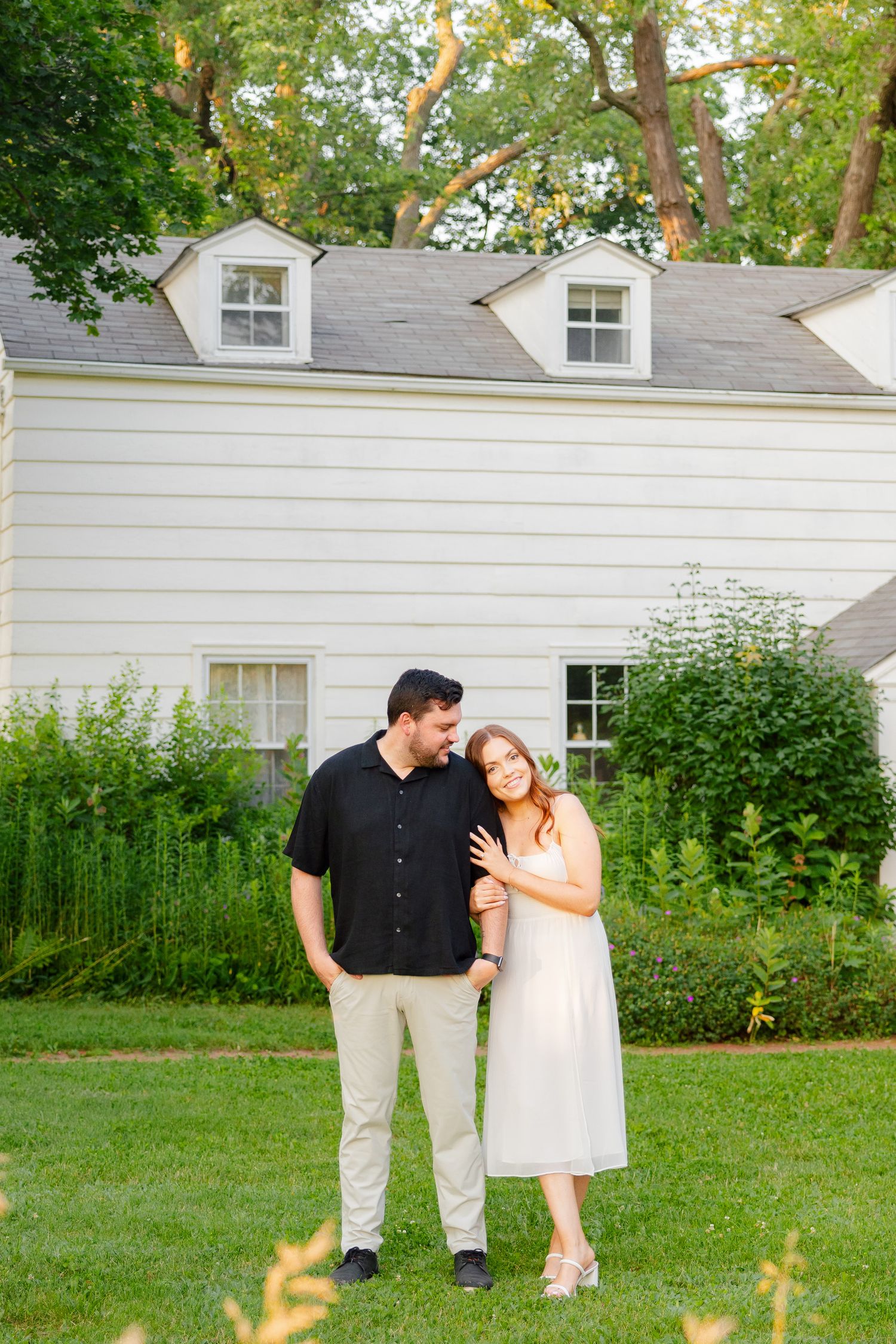 Tender moment between couple in garden setting with white colonial house behind them.