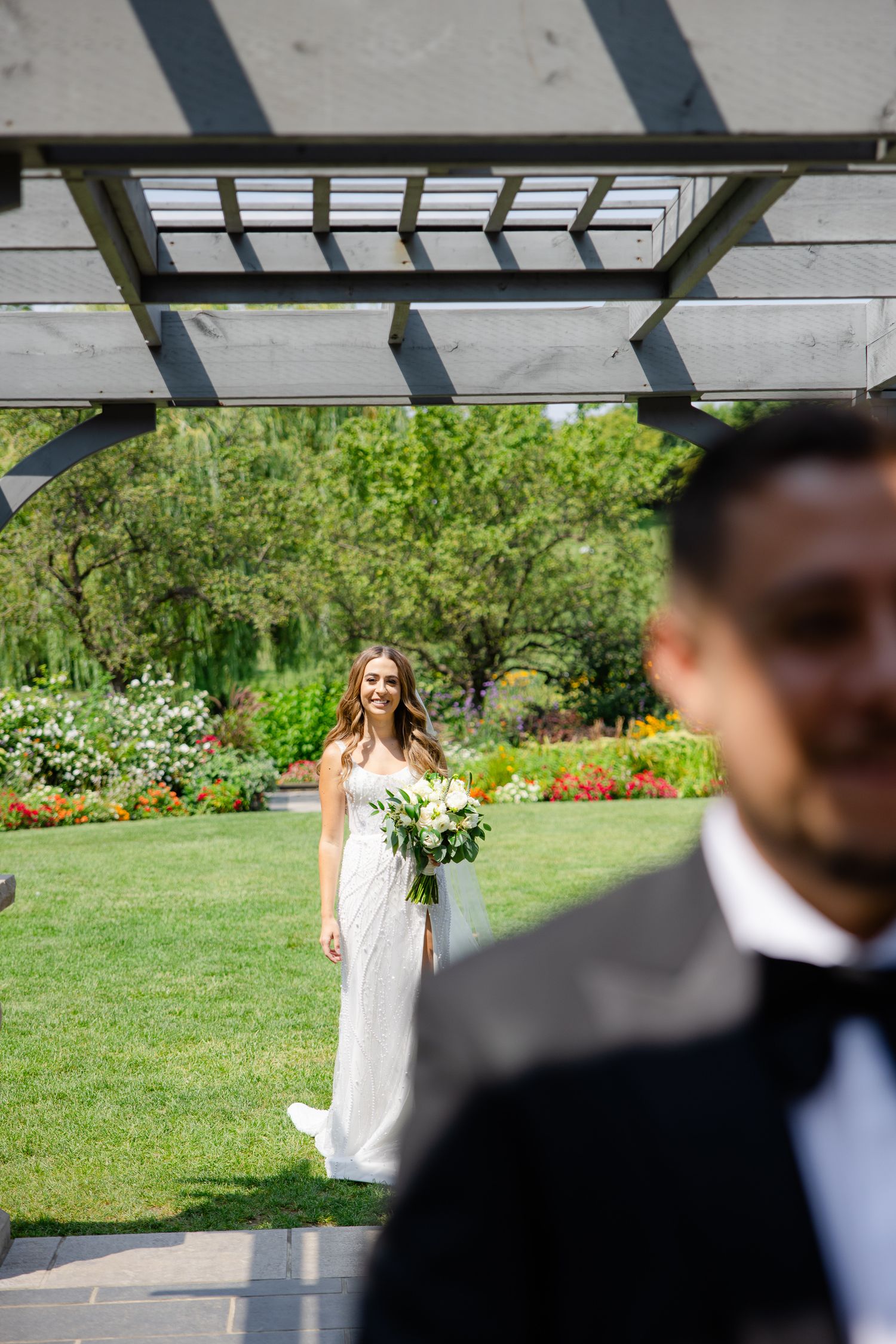A garden wedding scene with a pergola casting shadows as a couple shares a first look moment.