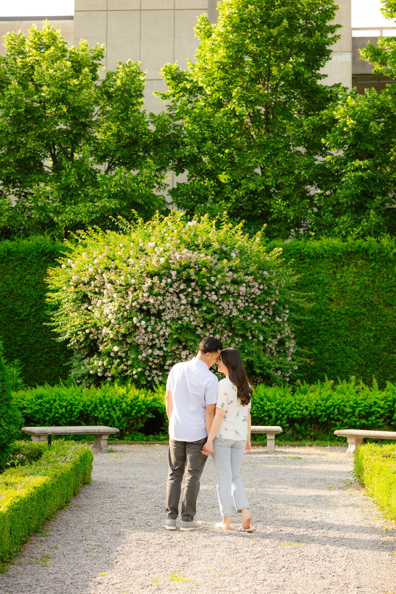 A couple shares an intimate moment on a garden path surrounded by flowering bushes and green trees.