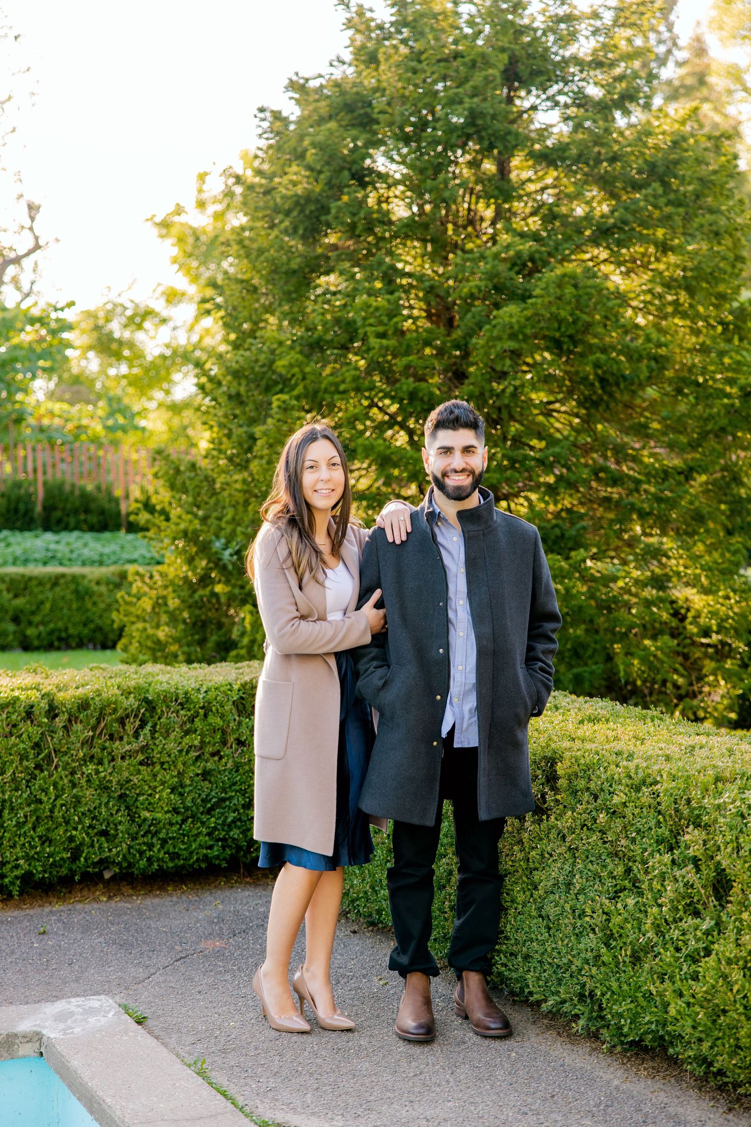 A couple stands together on a pathway surrounded by well-maintained hedges and trees.
