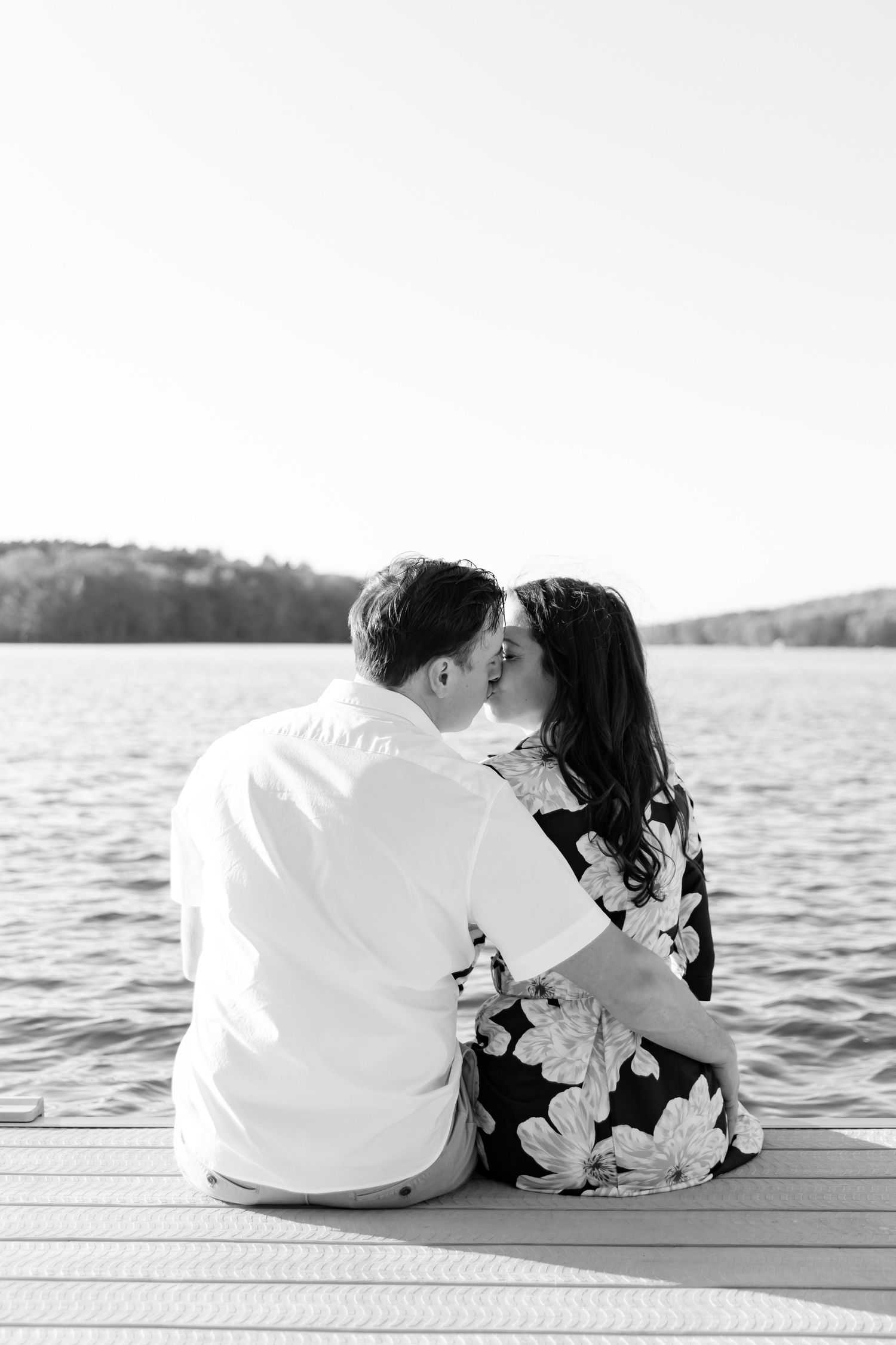 Black and white portrait of couple sitting close together on wooden dock by lake.