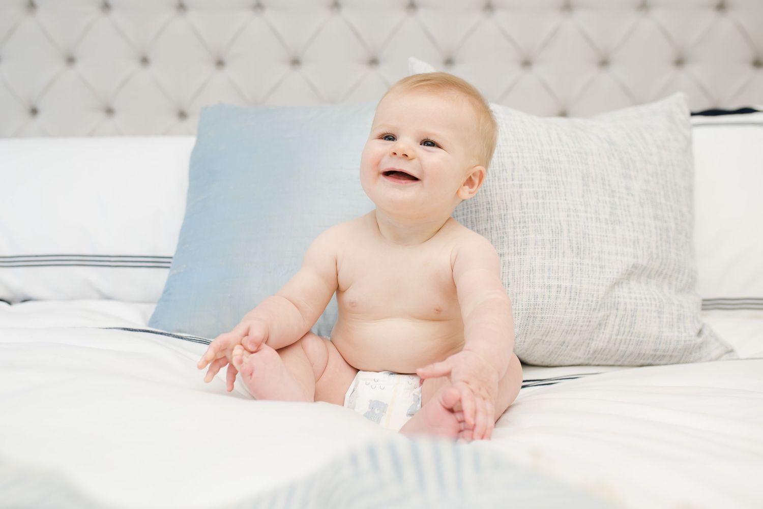 Baby in diaper sits smiling on white bedding with blue pillow.