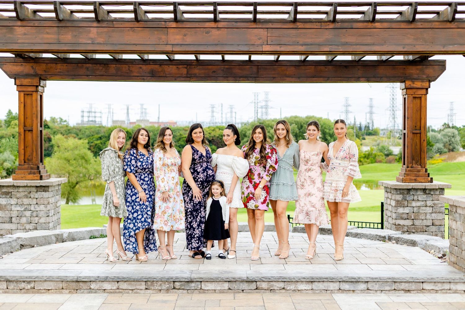 Group photo of guests in floral dresses standing under a wooden pergola on a stone patio with greenery in background.