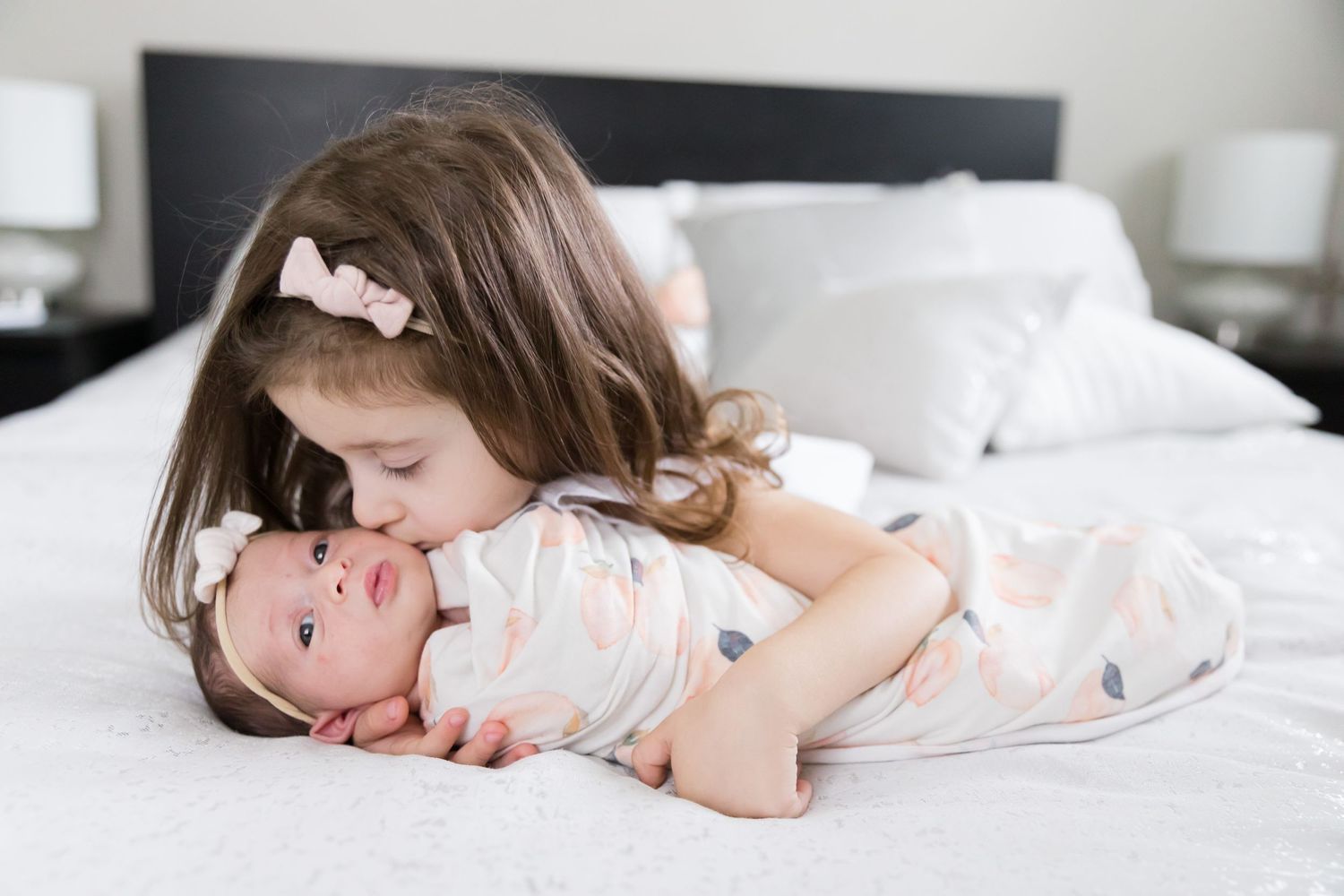 Older sibling lovingly embraces newborn baby on white bedding.